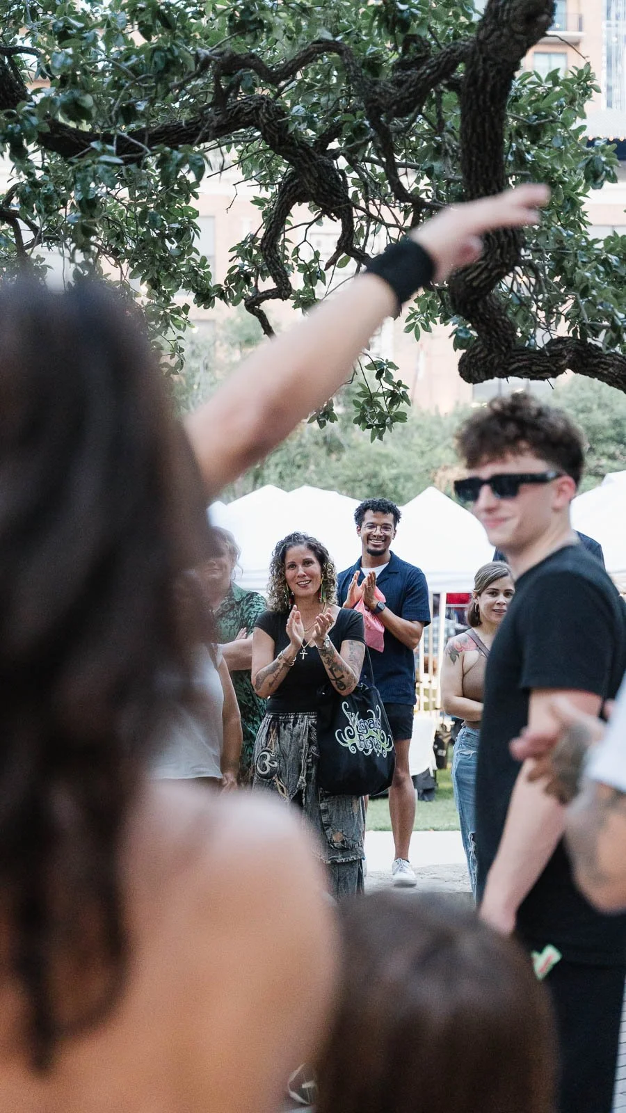 A group of diverse people gathered outdoors, some clapping and smiling, with tents and a large tree in the background.