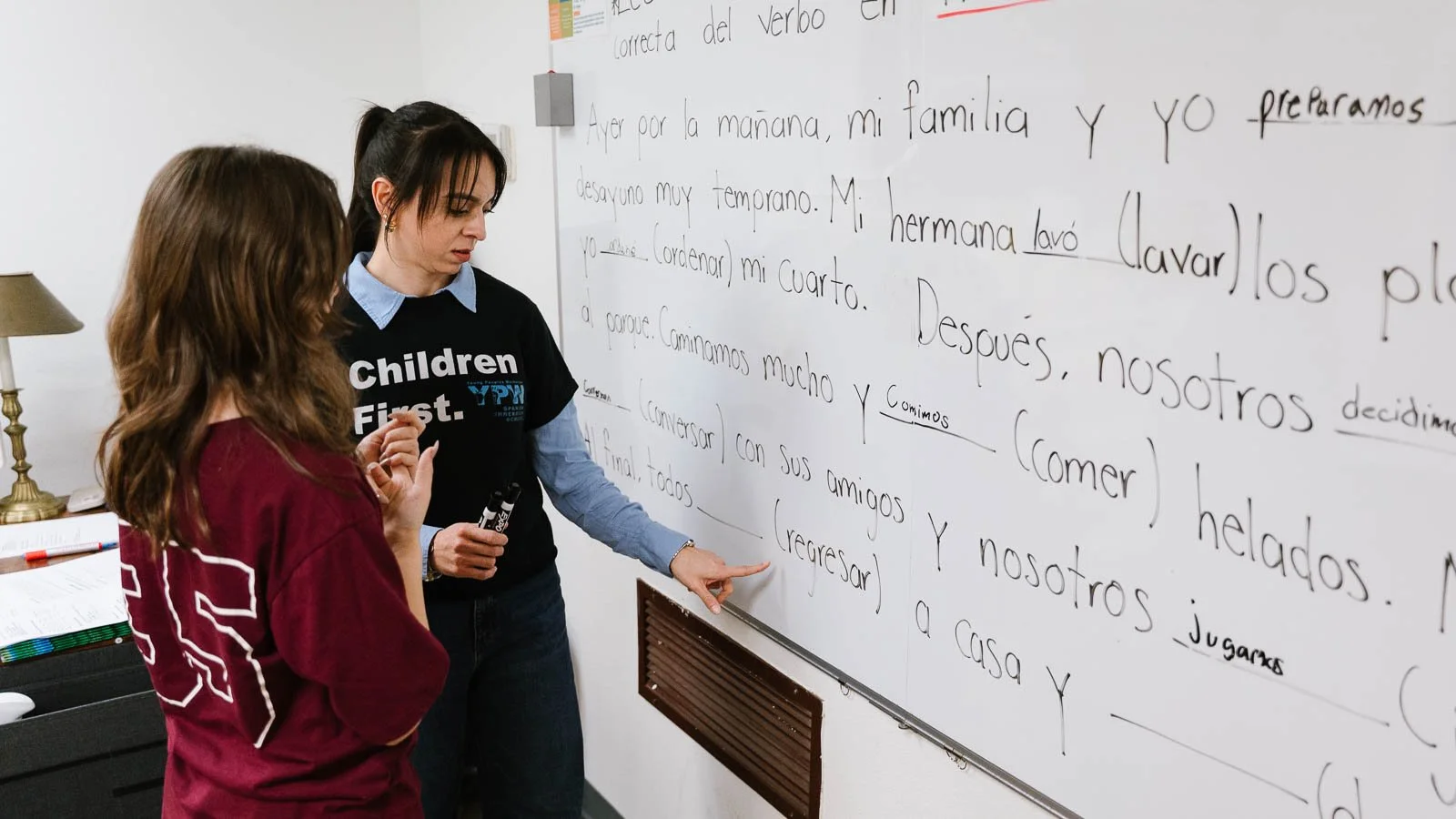 A teacher explains a Spanish worksheet to a student in a classroom. The worksheet has handwritten sentences in Spanish, and the teacher points at the writing with a marker. The classroom has a whiteboard and a music stand with colorful markers.