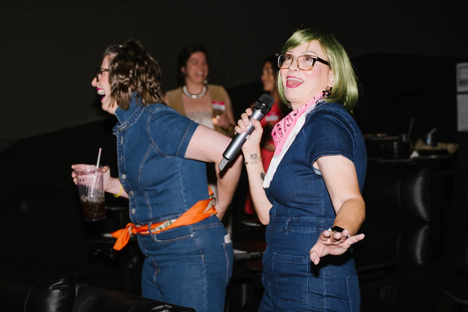 A woman with green hair and glasses singing into a microphone at a social gathering, others laughing and holding drinks in the background.