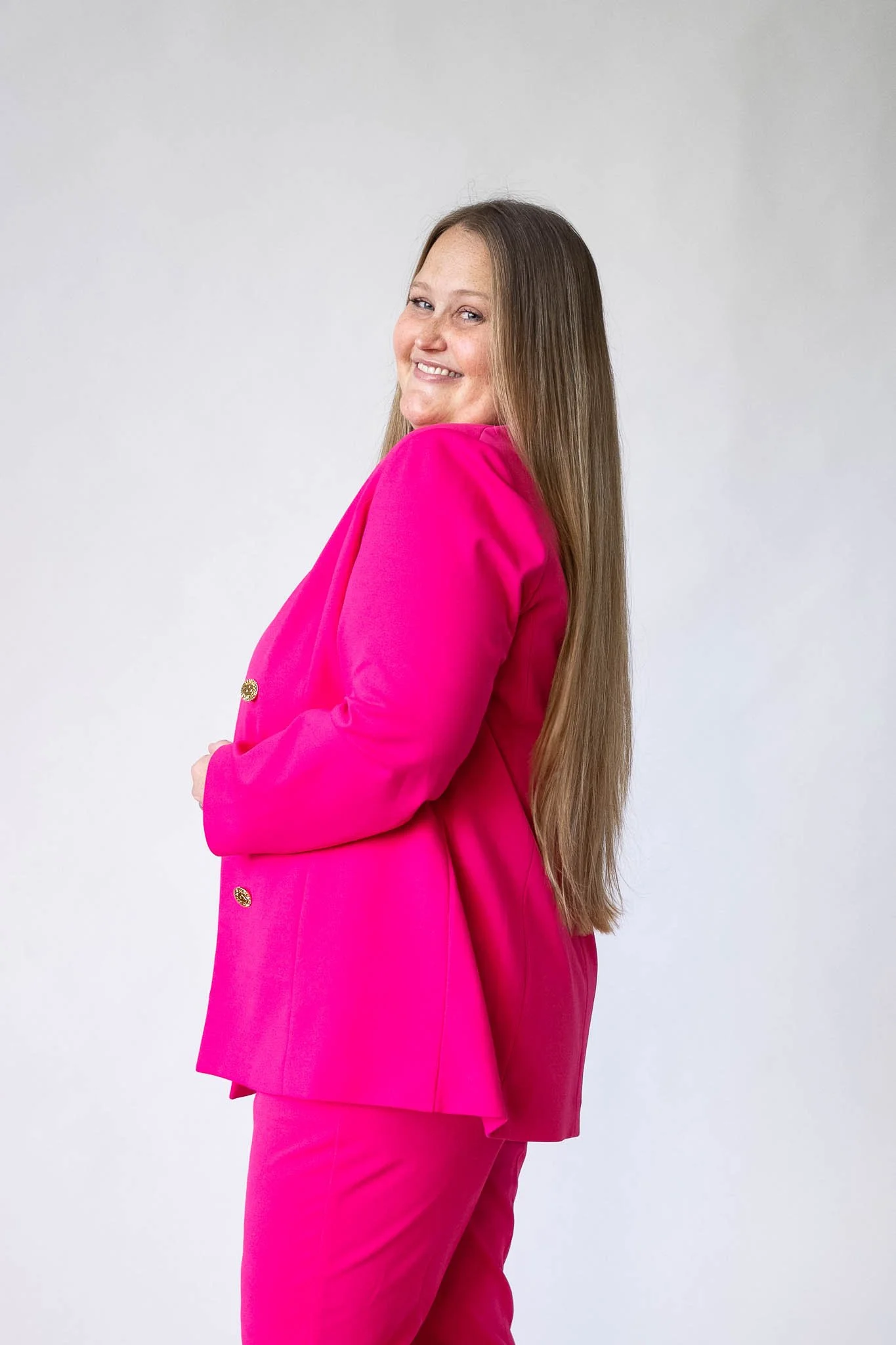 A smiling woman with long brown hair wearing a bright pink blazer and matching pants, standing against a plain white background.