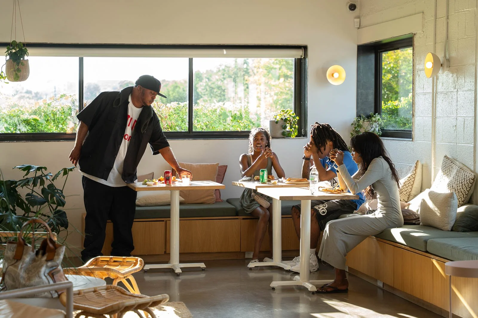 A group of four people and a person standing at a table in a bright, modern room with large windows and outdoor greenery background. They are eating and socializing.