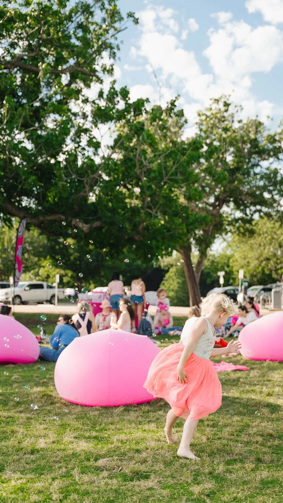 Children playing outdoors with pink inflatable pools, bubbles, and trees on a sunny day.