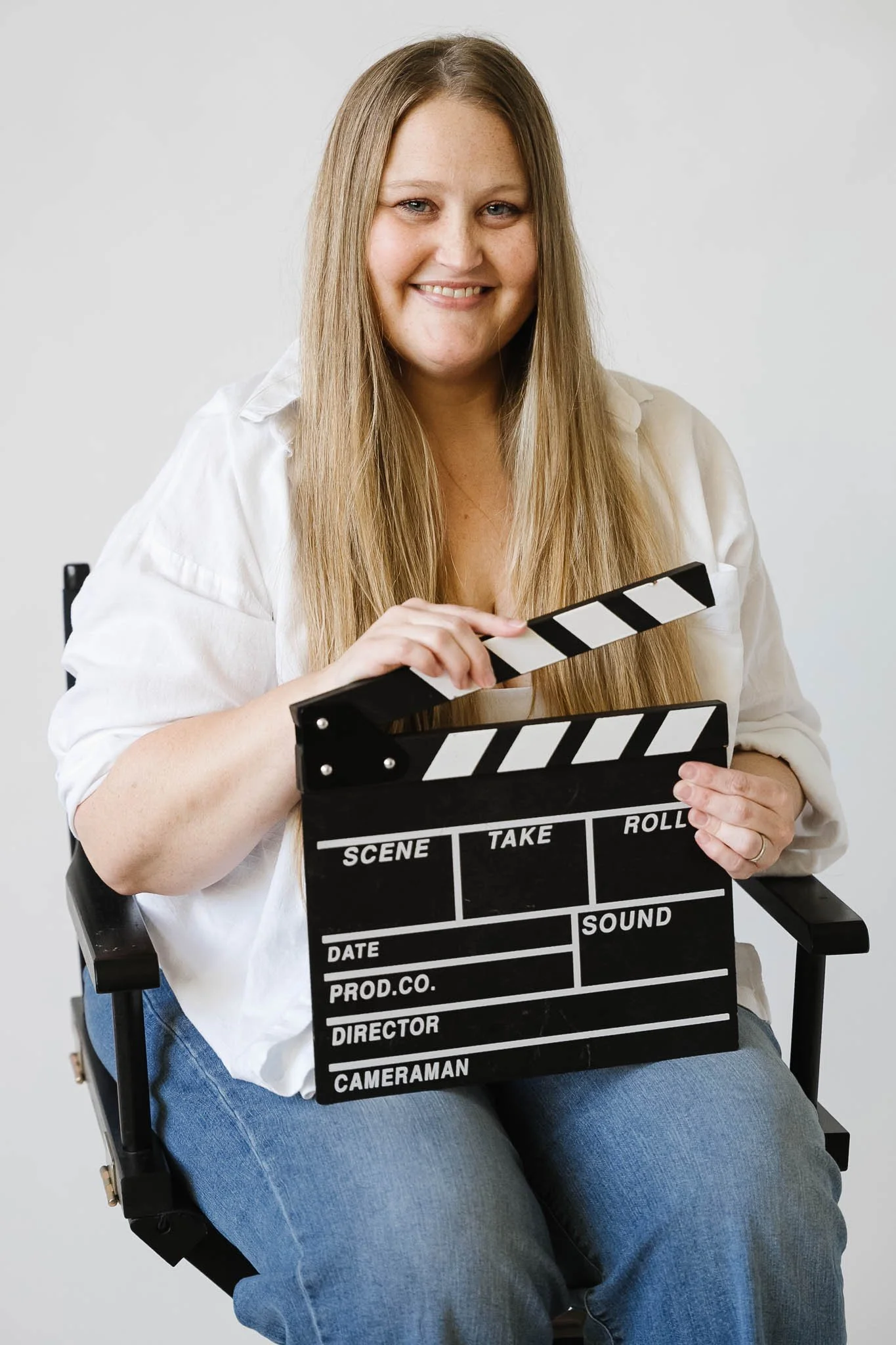 A woman smiling while sitting on a director's chair, holding a film slate. She has long, light brown hair, fair skin, and is wearing a white shirt and blue jeans against a plain light background.