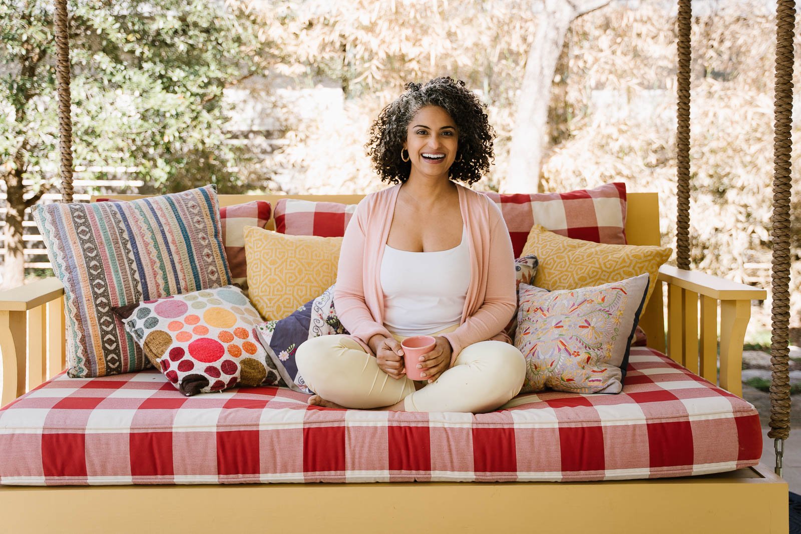 A woman with curly dark hair, wearing a light pink cardigan and cream pants, sitting cross-legged on a daybed with colorful pillows, holding a pink mug, outdoors with trees in the background.