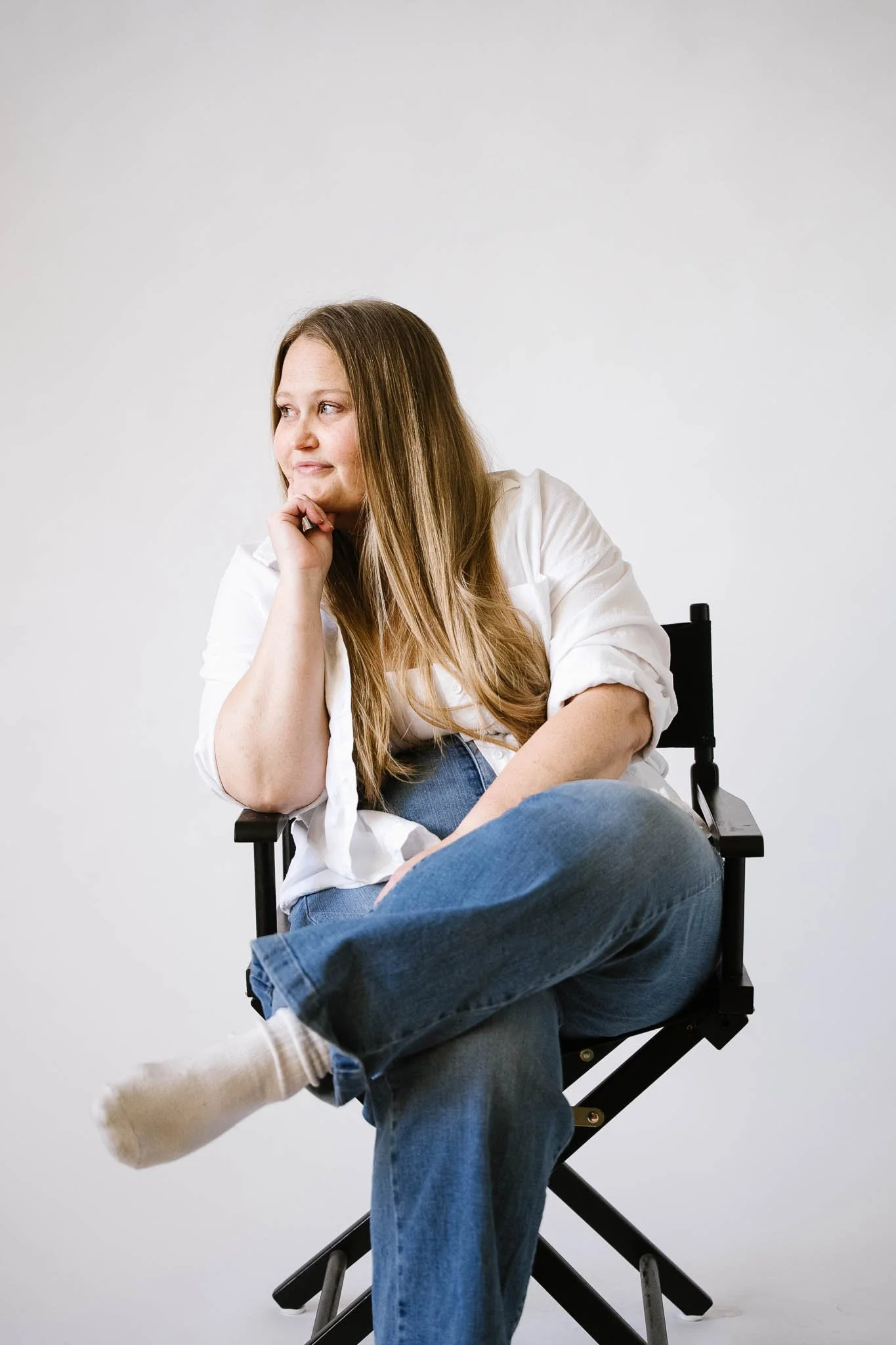 A woman with long hair sitting on a director's chair against a plain white background, wearing a white shirt and blue jeans, with her left leg crossed over her right, looking to her left with her chin resting on her left hand.