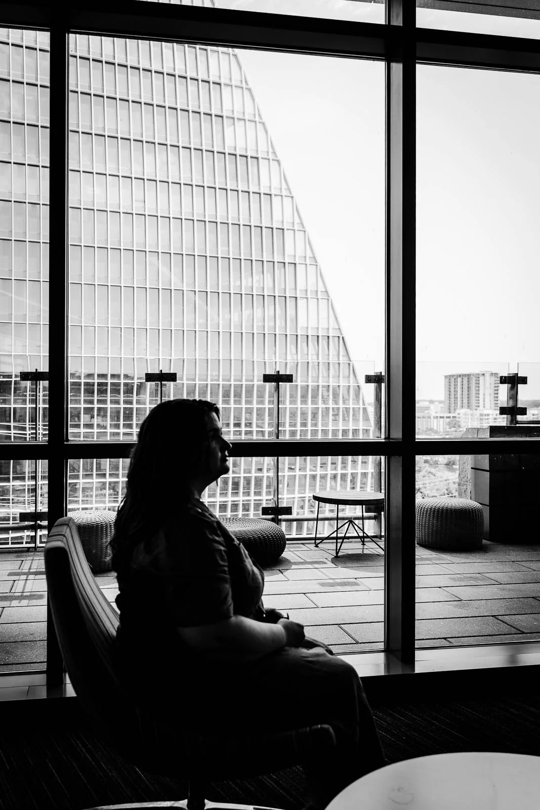 Silhouetted woman sitting on a chair inside a modern building, looking out a large window at cityscape and tall glass building.