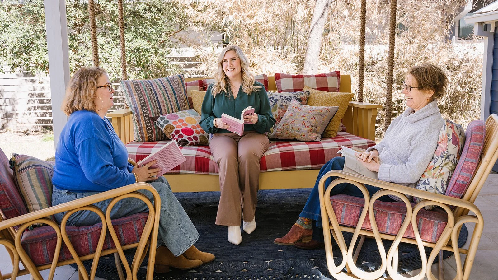 Three women sit on a porch with cushions, engaged in a book club discussion, holding books and smiling.