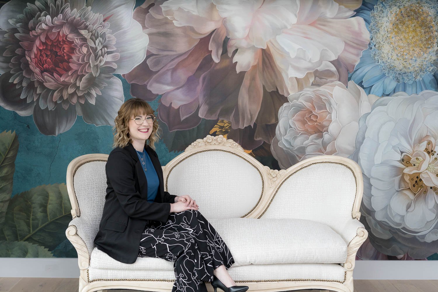 A woman with glasses and shoulder-length curly hair sitting on a vintage white sofa with a floral mural background.