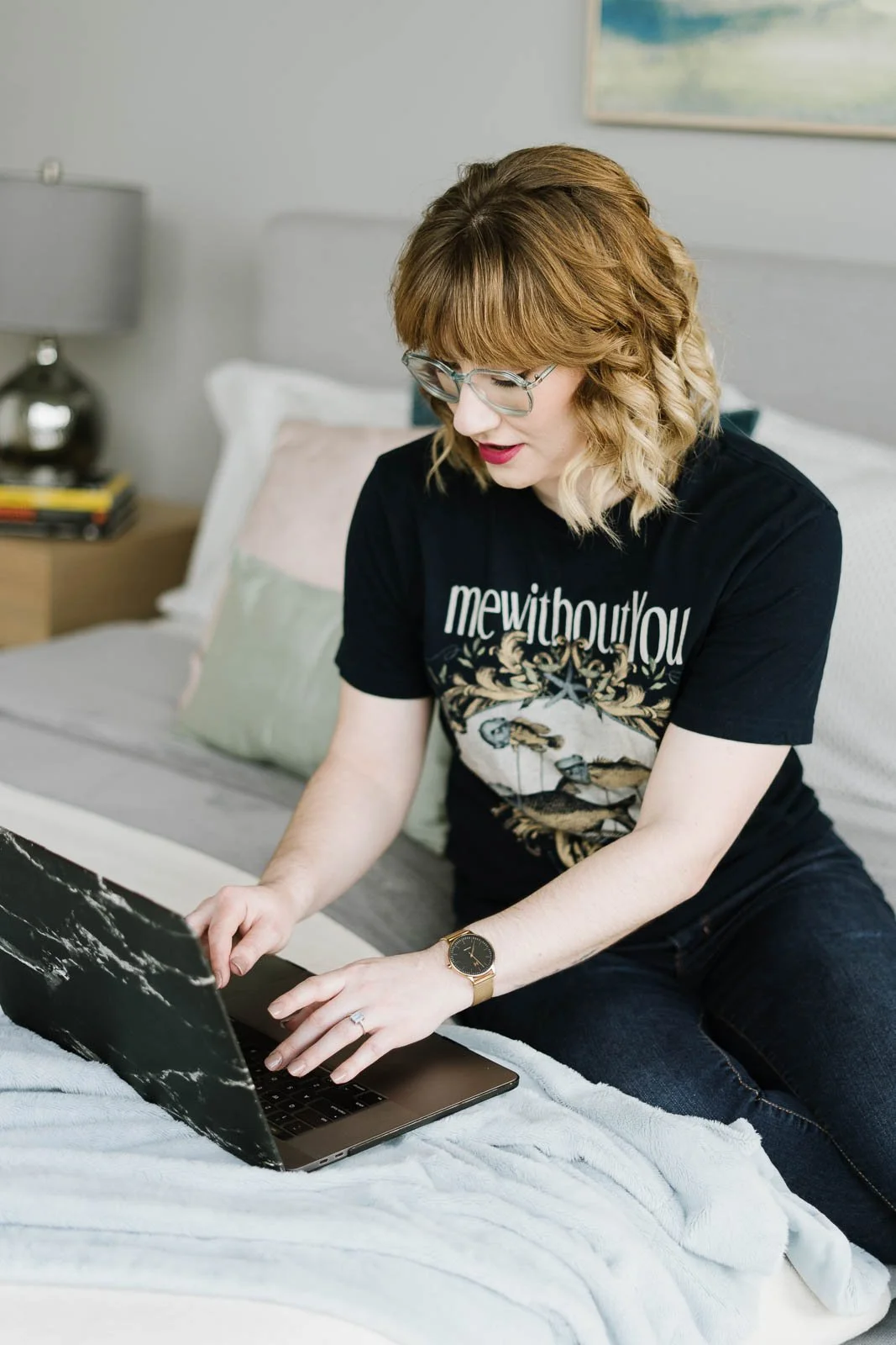 A woman with curly hair, glasses, and a black graphic t-shirt sits on a bed and uses a laptop.