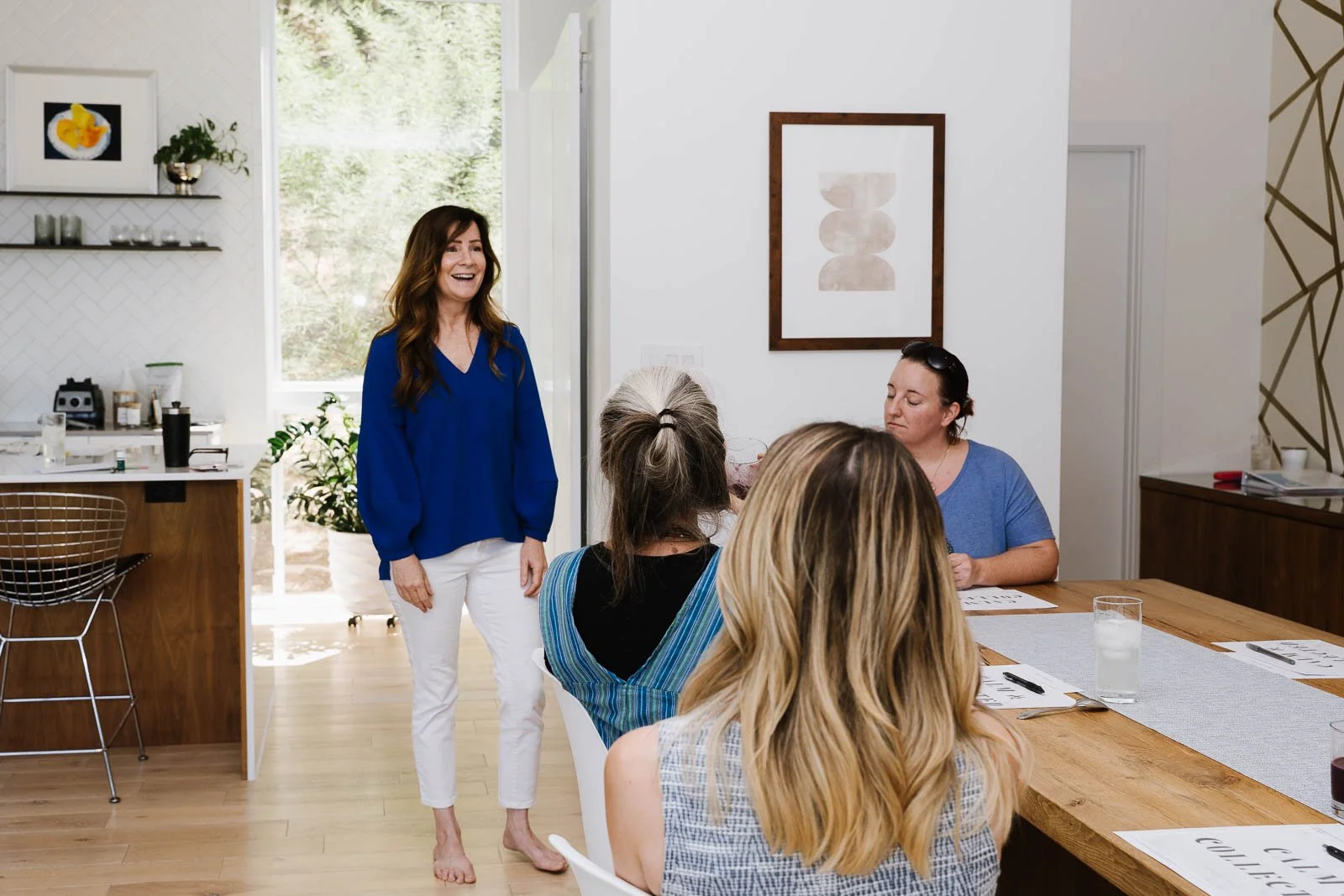 Five women in a meeting room, four seated at a table and one standing, smiling, wearing a blue top. The room has white and wood decor, with a large window and natural light.