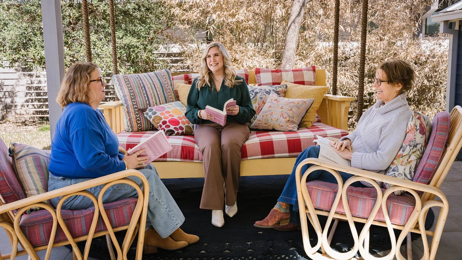 Three women sitting outdoors on chairs and a couch, reading books and chatting on a porch with trees in the background.