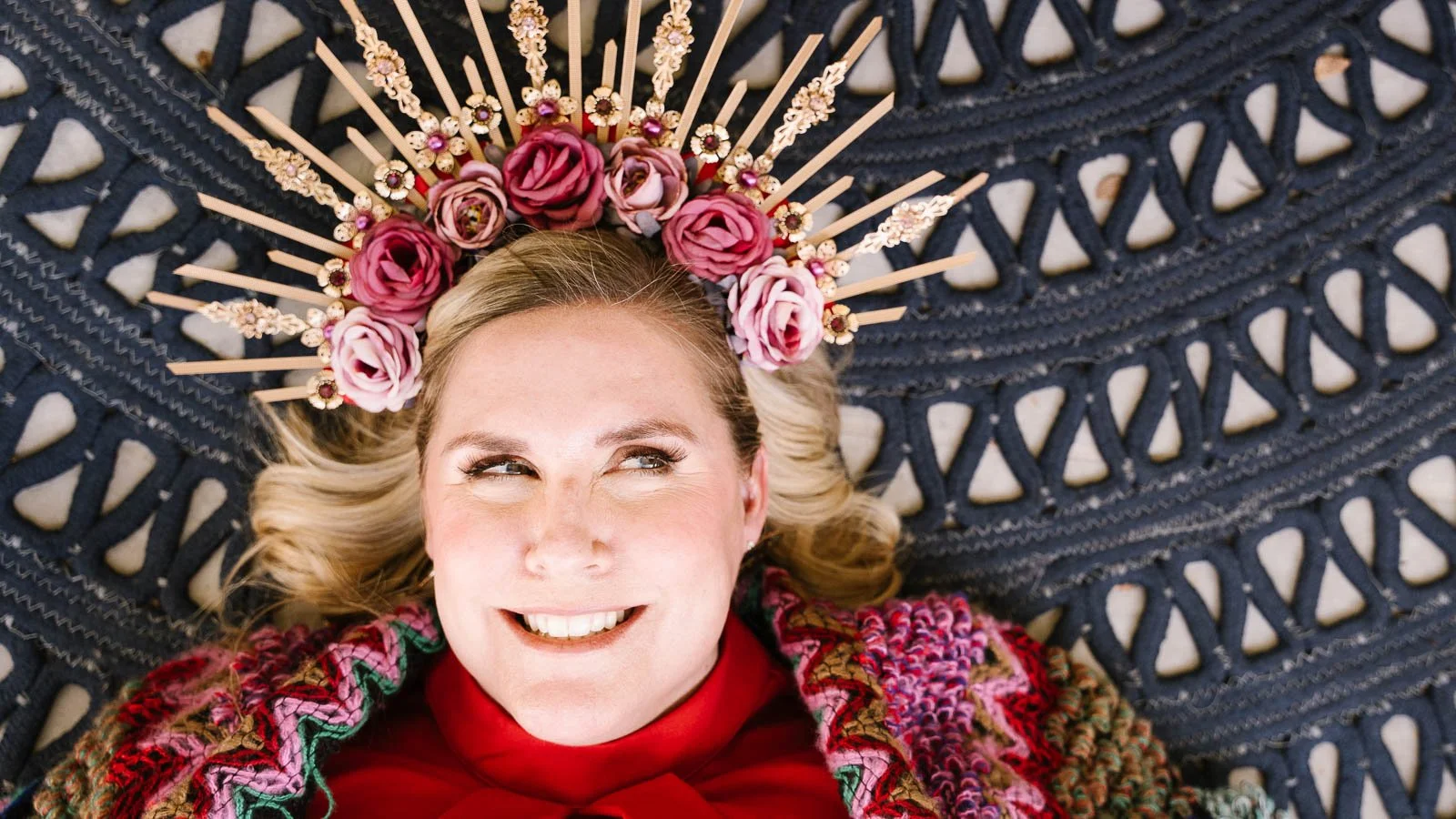 A woman with blonde hair lying down on a textured navy blue surface, smiling and looking to the side. She is wearing a headpiece with pink roses and gold embellishments, and a multicolored knitted garment with red and pink hues.