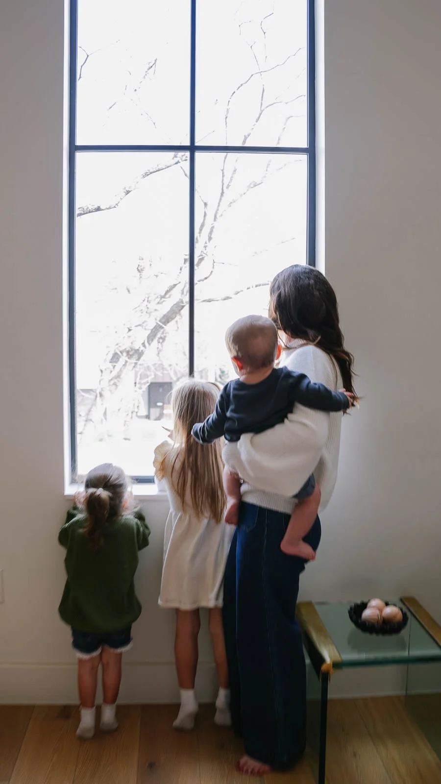 A woman with four children looking out a large window during winter, with snow and a tree outside.
