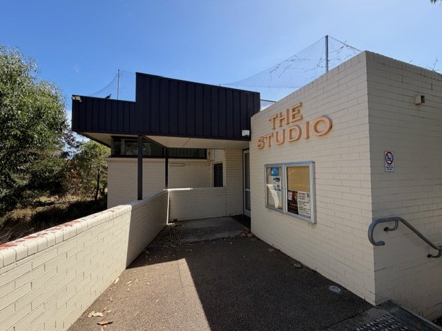 Exterior of a building labeled 'The Studio' with a small window and a bulletin board outside, surrounded by trees and a clear blue sky.
