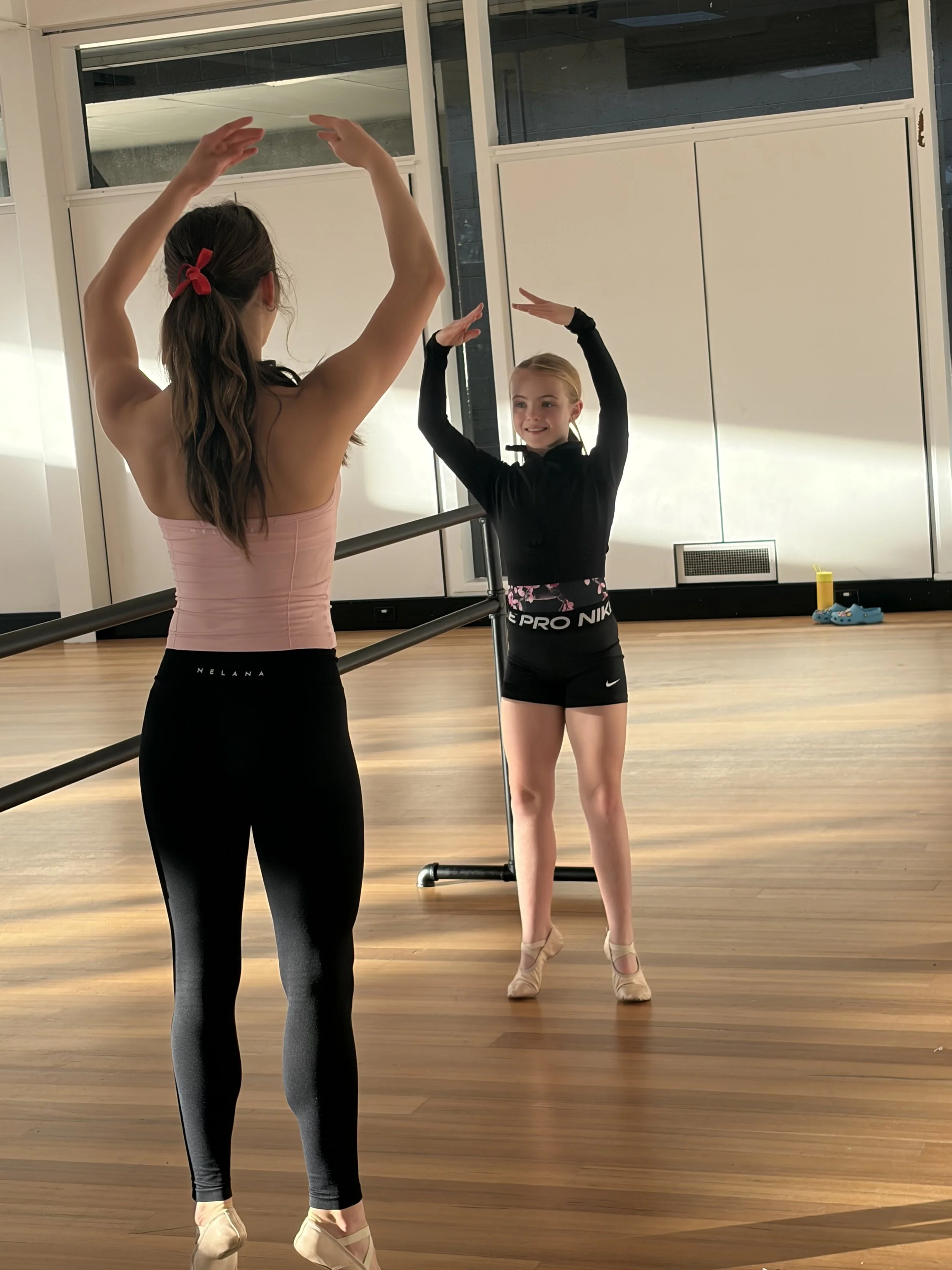 A ballet instructor in black leggings and a pink tank top demonstrates a ballet pose for a young girl in black dance practice attire in a dance studio with wood flooring and mirrored wall.