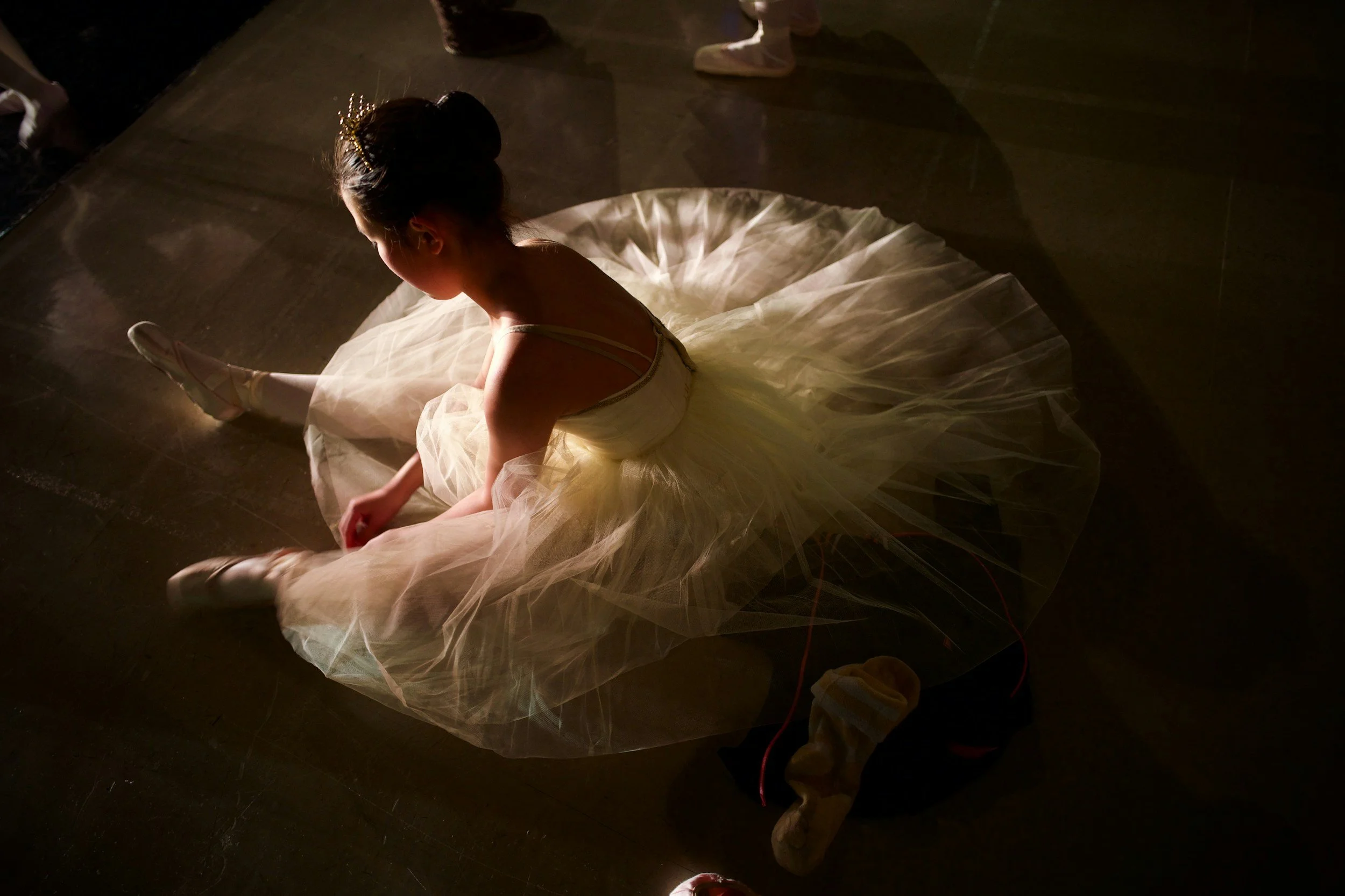 A young ballet dancer sitting on the dark floor with her legs extended forward. She is wearing a light-colored tutu and ballet slippers, with her hair styled in a bun adorned with a decorative headpiece. The lighting creates a dramatic shadow on the floor.
