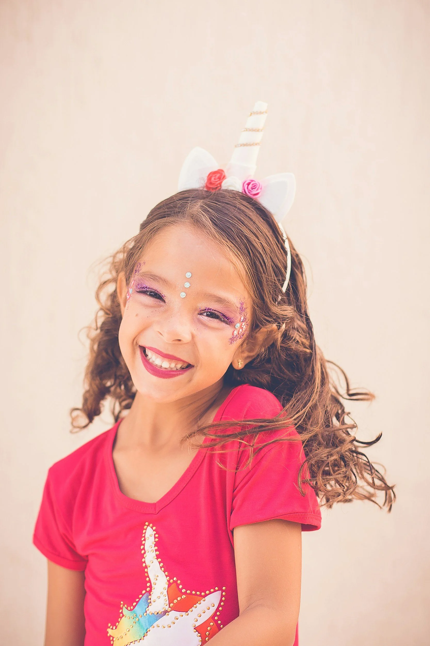 A young girl wearing a unicorn headband, smiling, with face glitter and rhinestones, dressed in a pink T-shirt with a unicorn design.