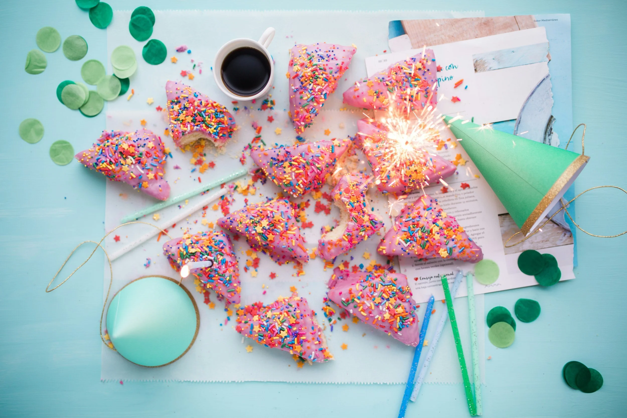 A birthday celebration with a half-eaten pink frosted cake decorated with colorful sprinkles and a sparkler, a cup of black coffee, green paper confetti, and party hats on a light blue table.