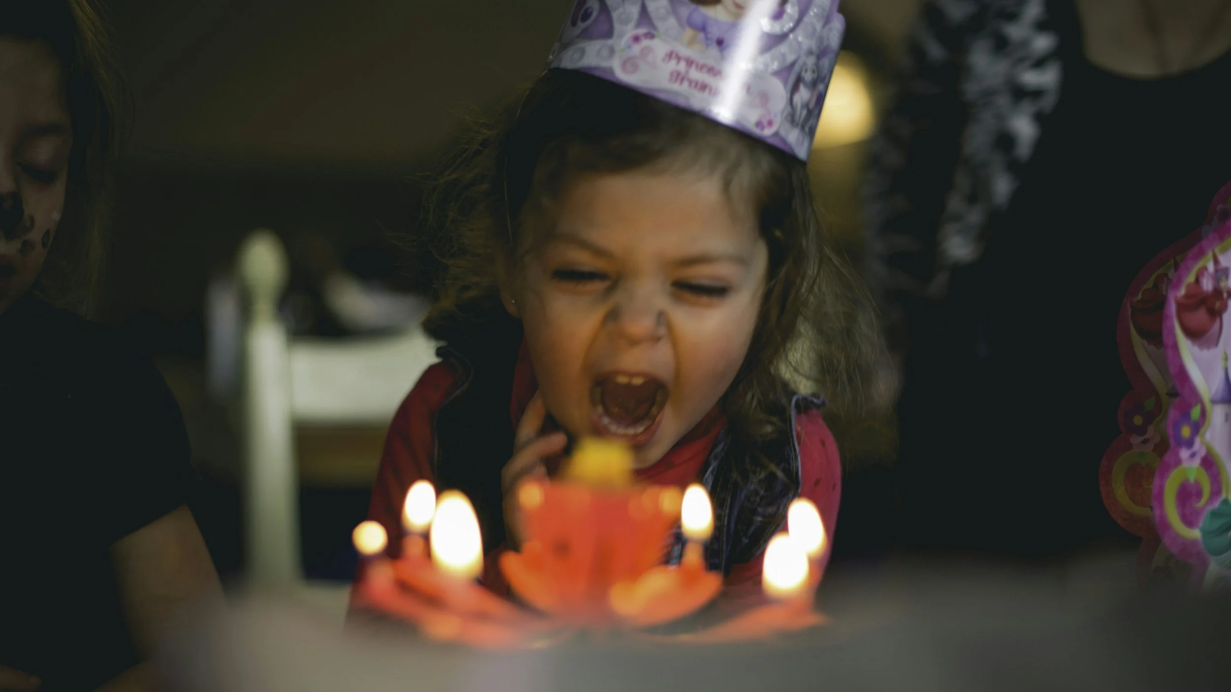 A young girl with curly hair wearing a birthday hat, celebrating her birthday with a yellow cake and candles, party decorations in the background.
