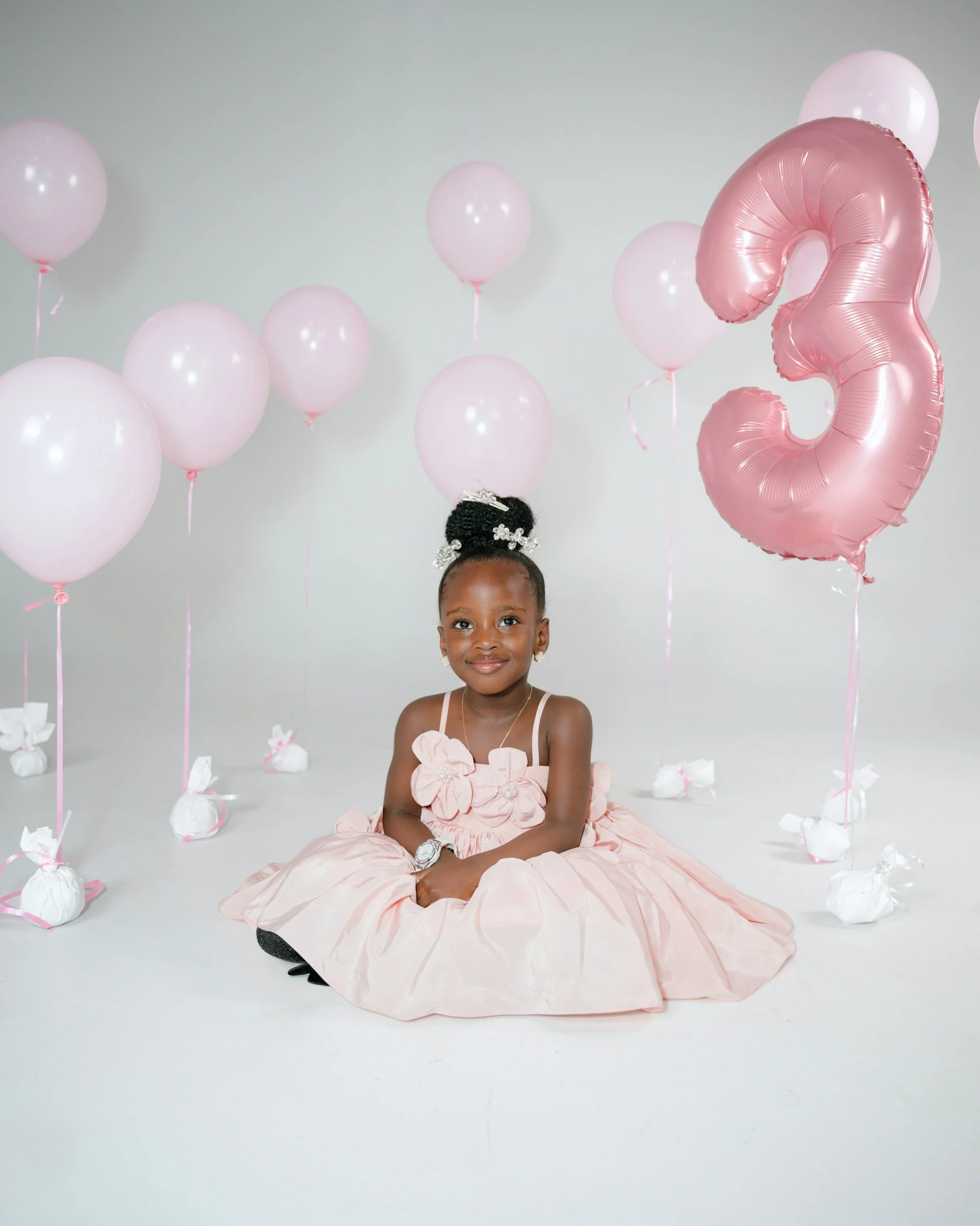 Young girl in a pink dress sitting on the floor with balloons around her, against a plain white background.