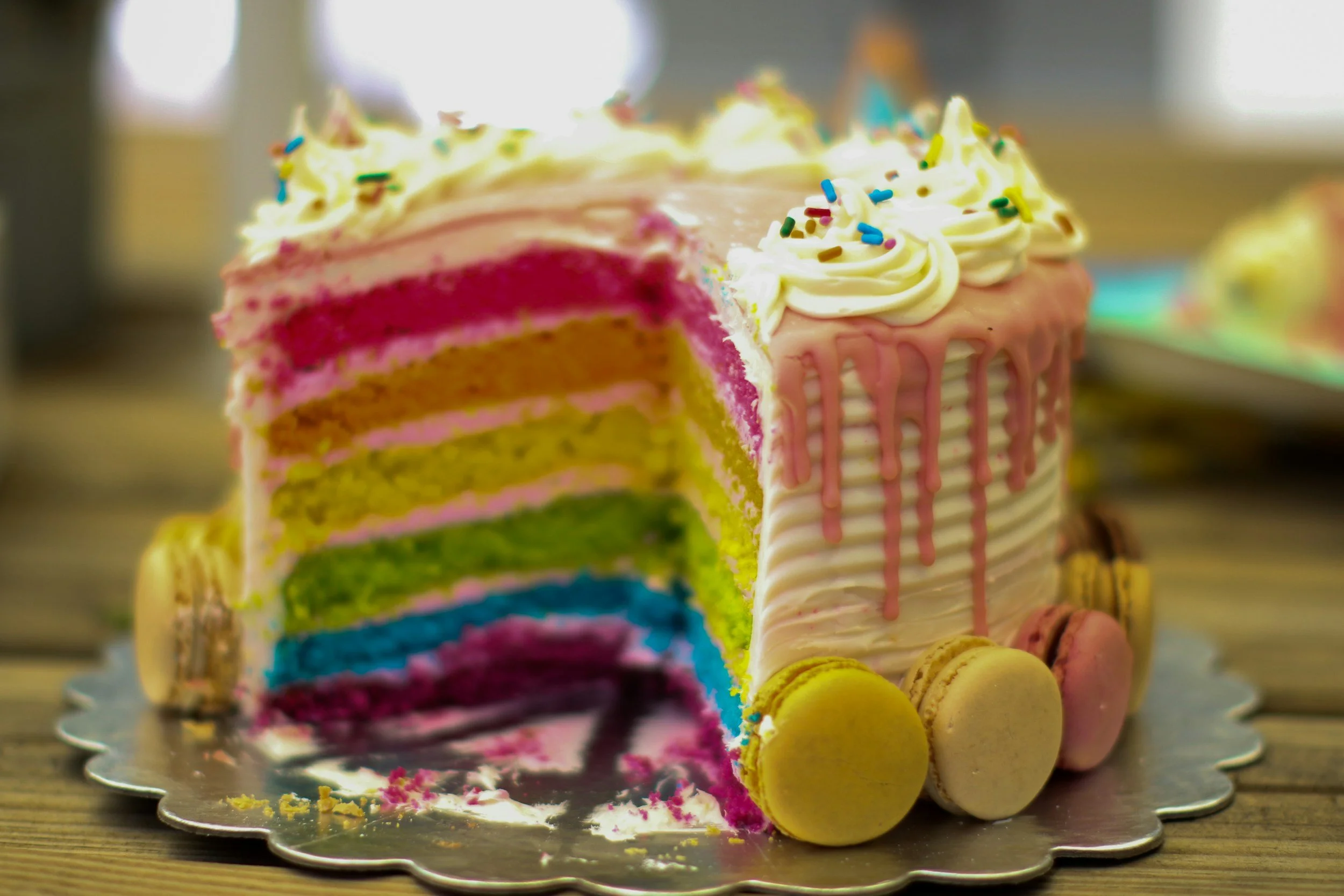 A partially sliced colorful, layered rainbow cake with white icing, sprinkles, and macarons on a cake board.