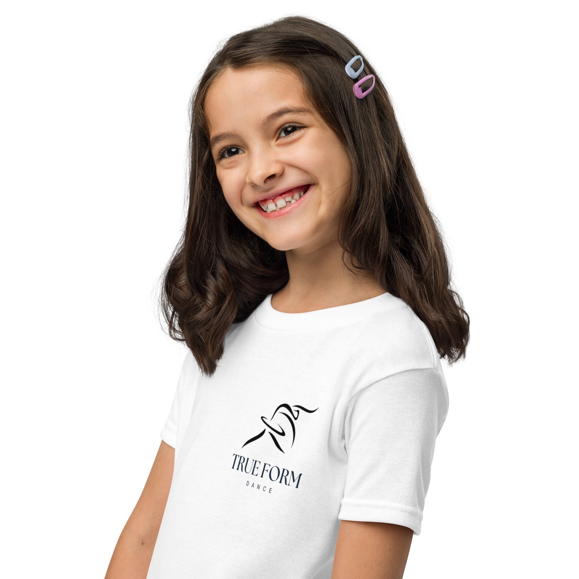 A young girl with long brown hair clipped with pastel-colored hair clips, smiling and showing her missing front teeth, wearing a white shirt with black text that says "TRUEFORM DANCE" on it.