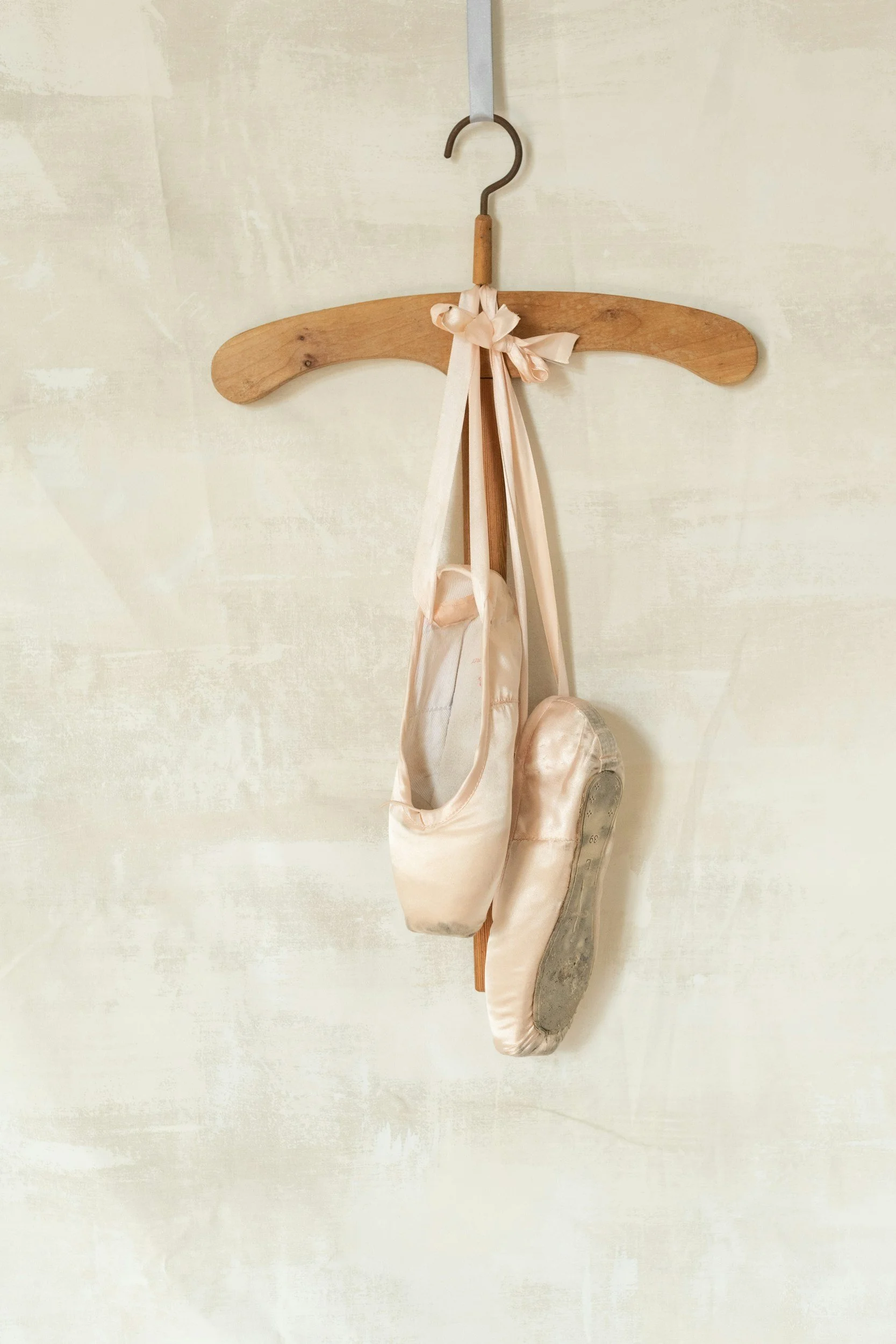 A pair of ballet pointe shoes hanging from a wooden hanger with a pink ribbon bow, mounted on a light-colored wall.