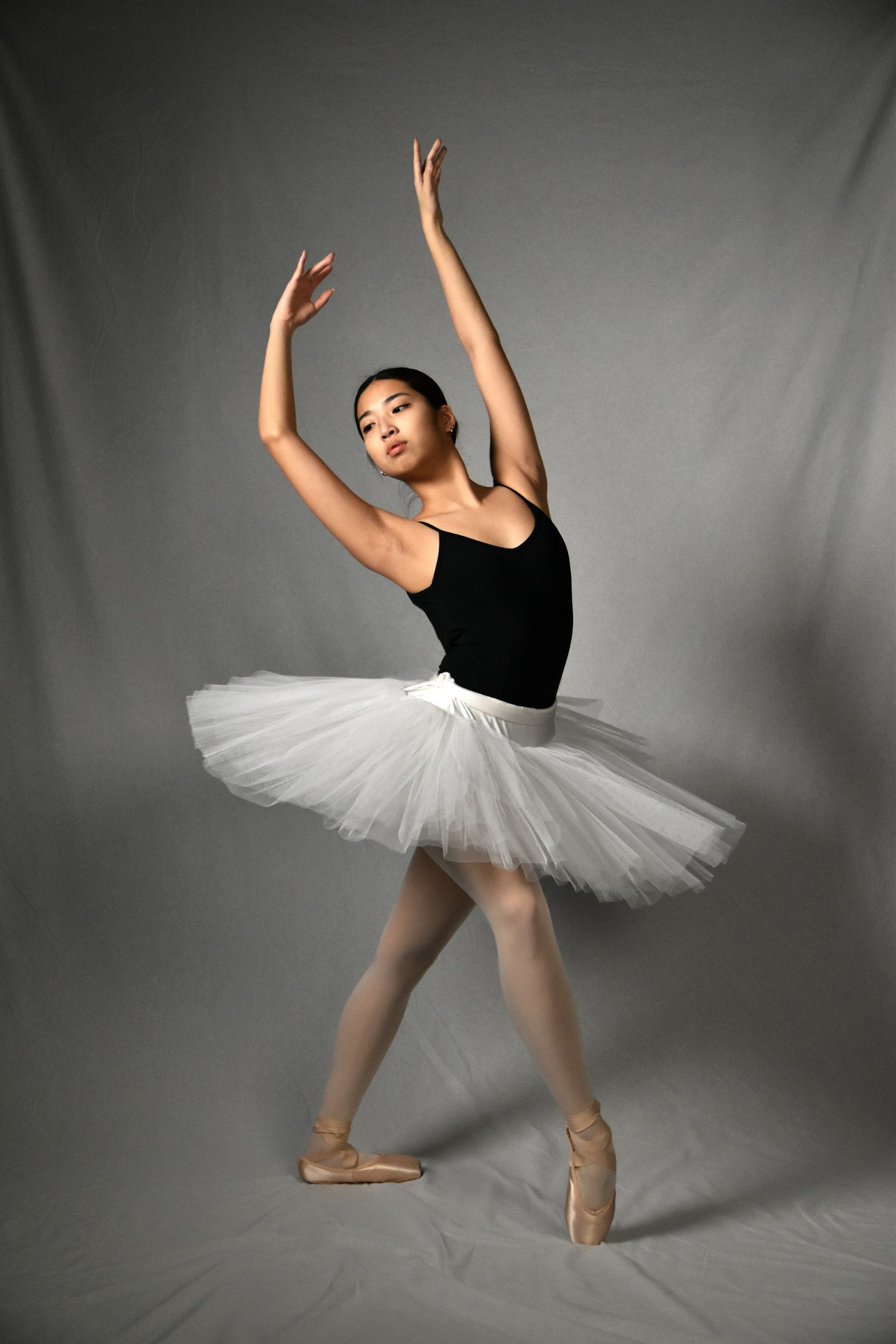 A ballet dancer in a black leotard and white tutu is performing on pointe, with one arm raised above her head and the other extended to the side, against a gray backdrop.