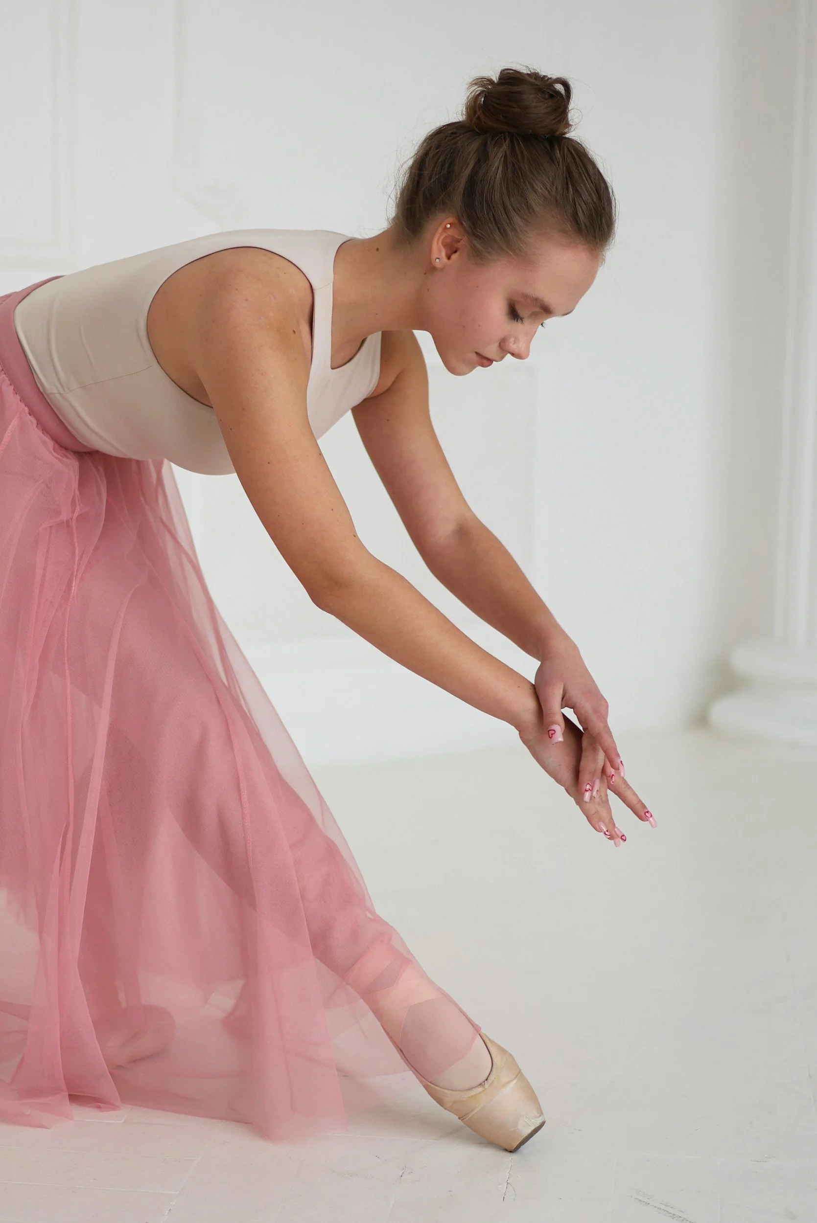 A young woman in a pink ballet tutu and pointe shoes is stretching or warming up in a white room with minimal decor.