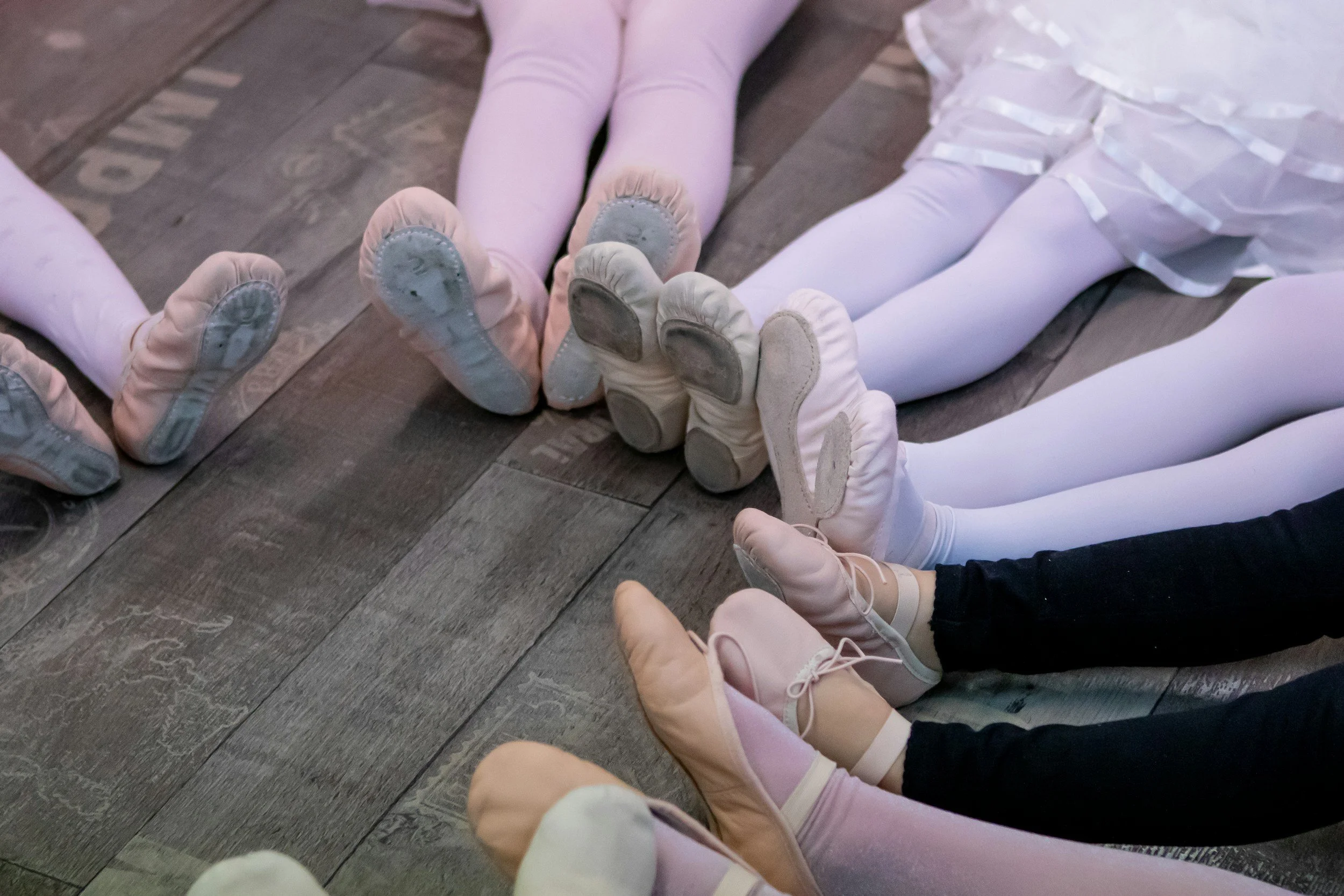 Children lying on a wooden floor, wearing ballet slippers and white tights, forming a circle with their legs and feet.