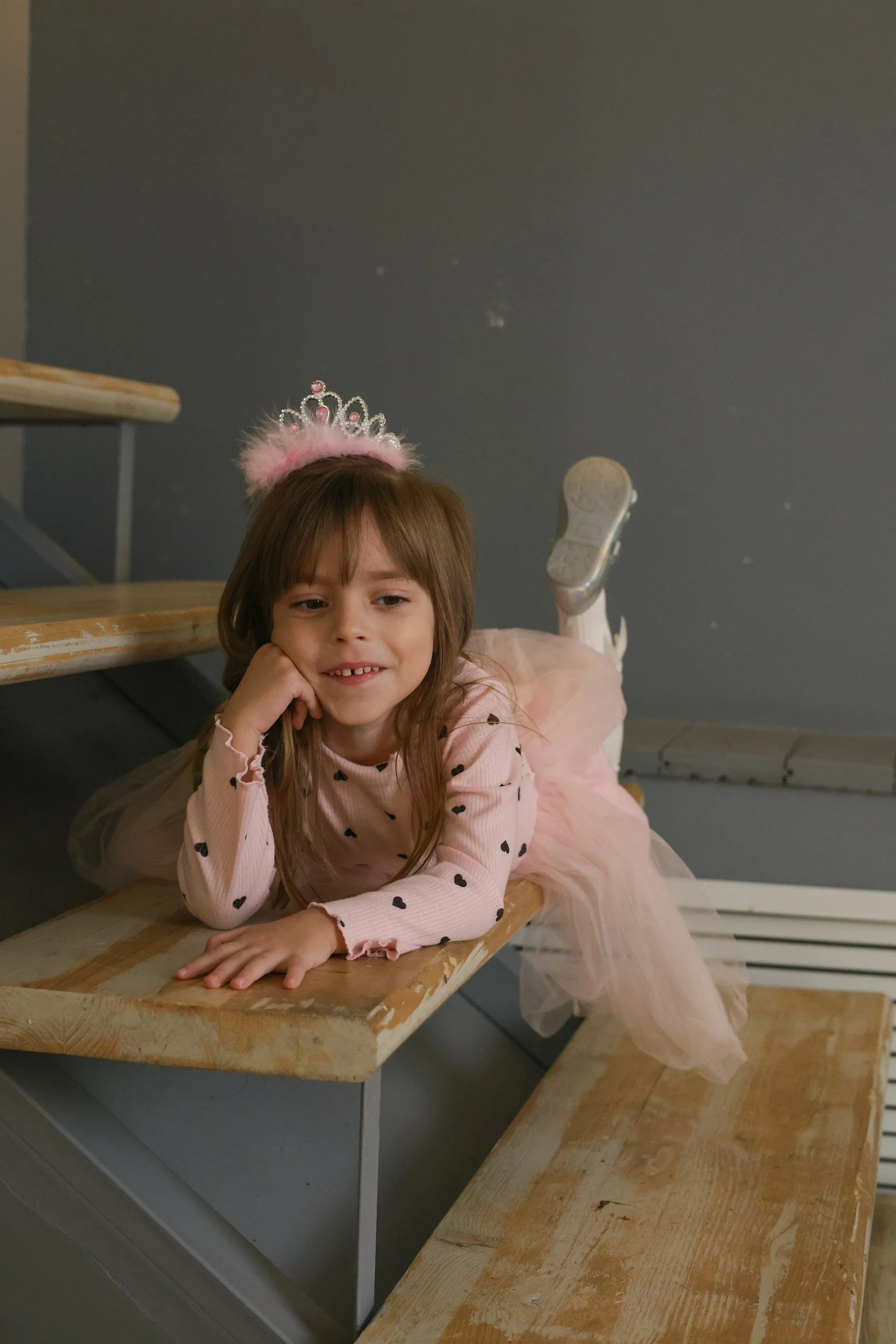 A young girl with brown hair and a pink dress, wearing a silver tiara with pink feathers, lying on a wooden staircase with a gray wall in the background.