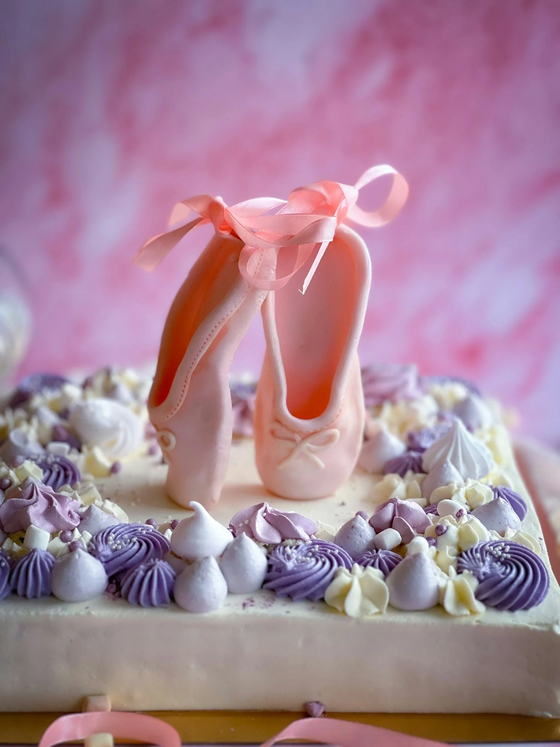 A ballet-themed cake decorated with pink ballet slippers on top and purple and white piped frosting borders.
