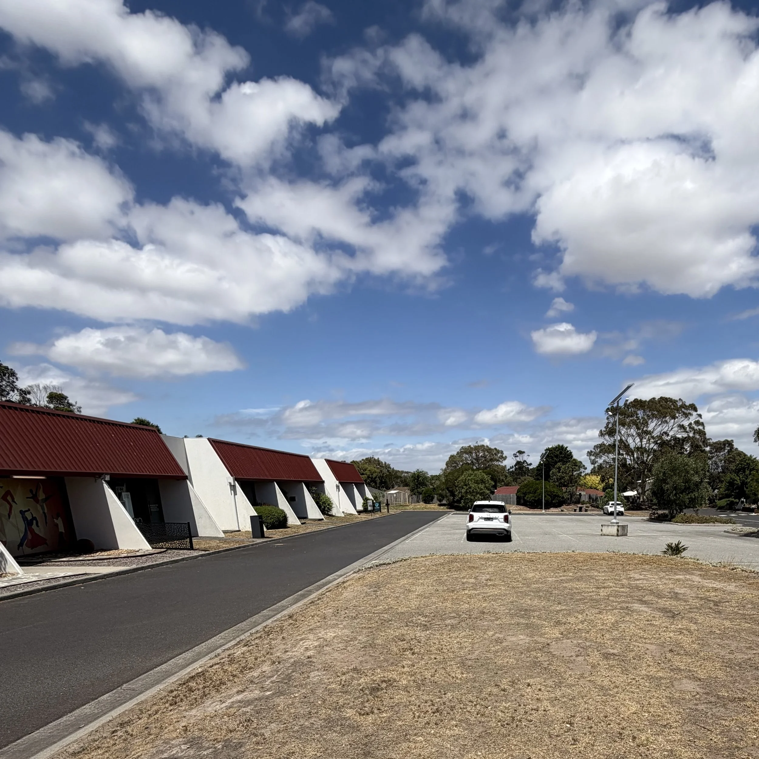 A parking lot with a white car parked near a curb, surrounded by trees and buildings, with a blue sky and scattered clouds overhead.