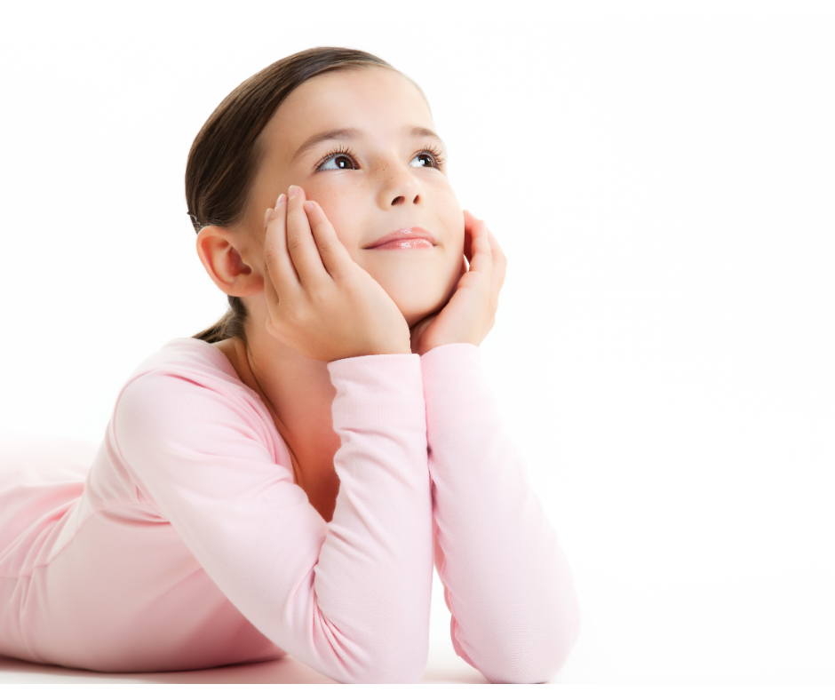A young girl lying on her stomach with her hands on her cheeks, looking upwards with a thoughtful expression, wearing a pink long-sleeve shirt.