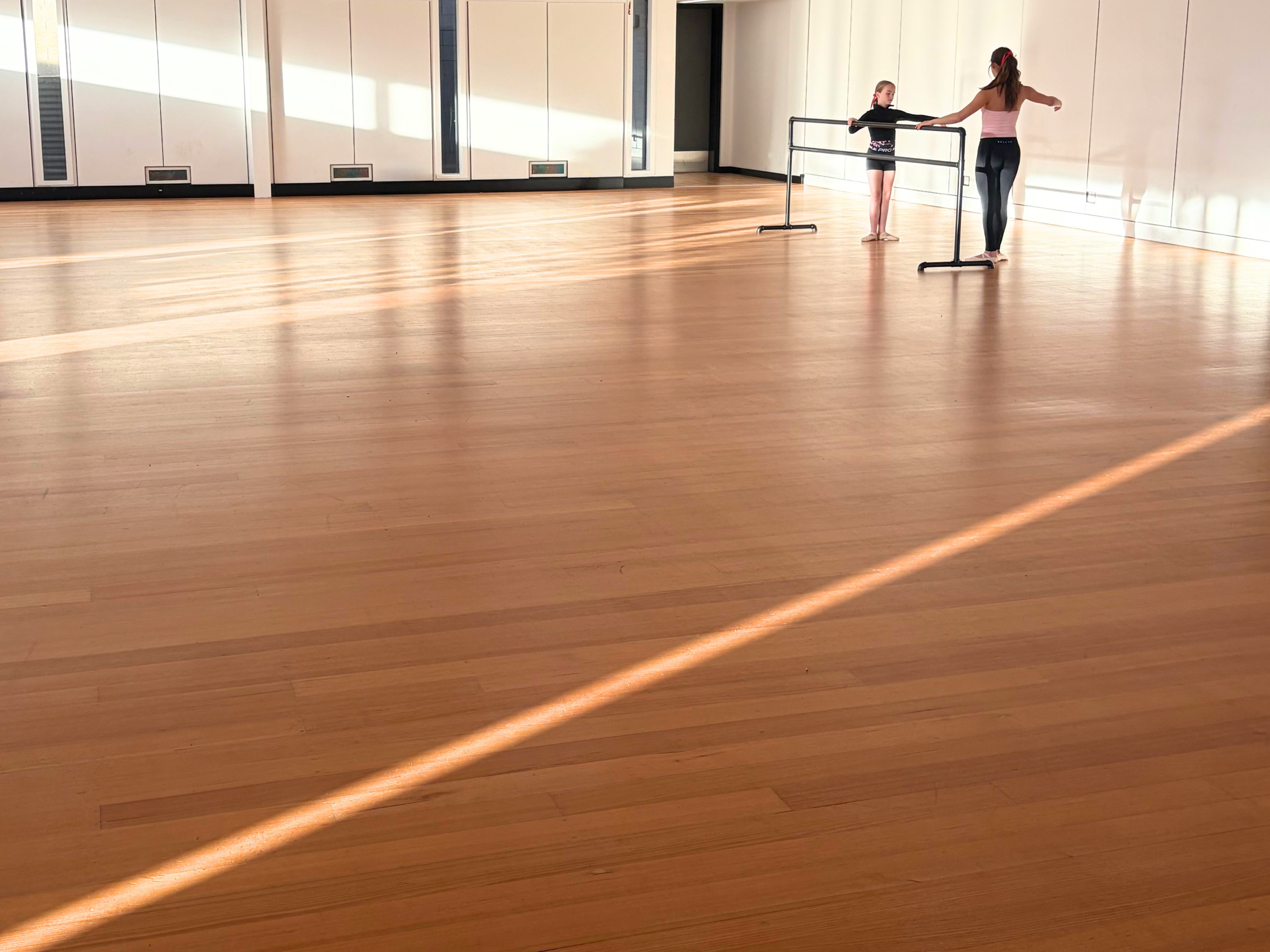 Two young girls practicing ballet at a dance studio with wooden floors and ballet barres.