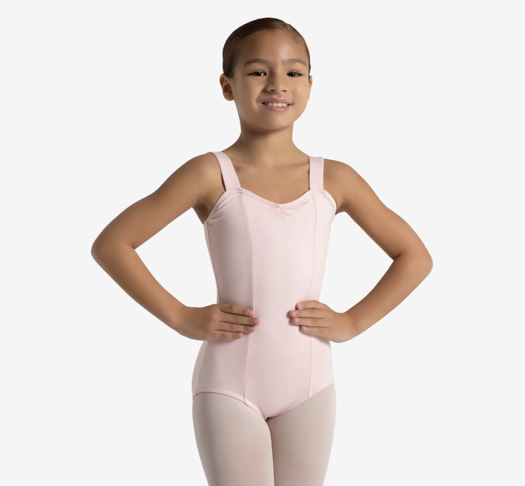 A young girl wearing a pink ballet leotard and tights, standing with hands on her hips, smiling against a plain white background.