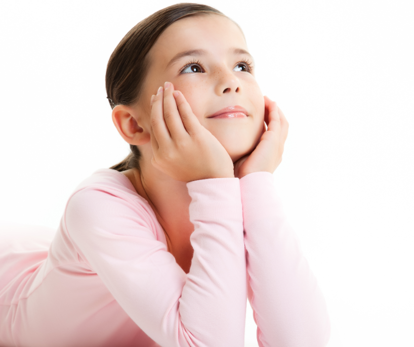 A young girl with brown hair, wearing a pink long-sleeve shirt, laying on her stomach with her hands on her cheeks, smiling and looking up.
