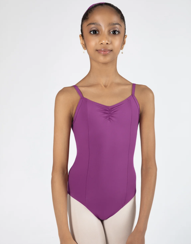 Young girl wearing a purple leotard and beige dance tights, standing against a white background.
