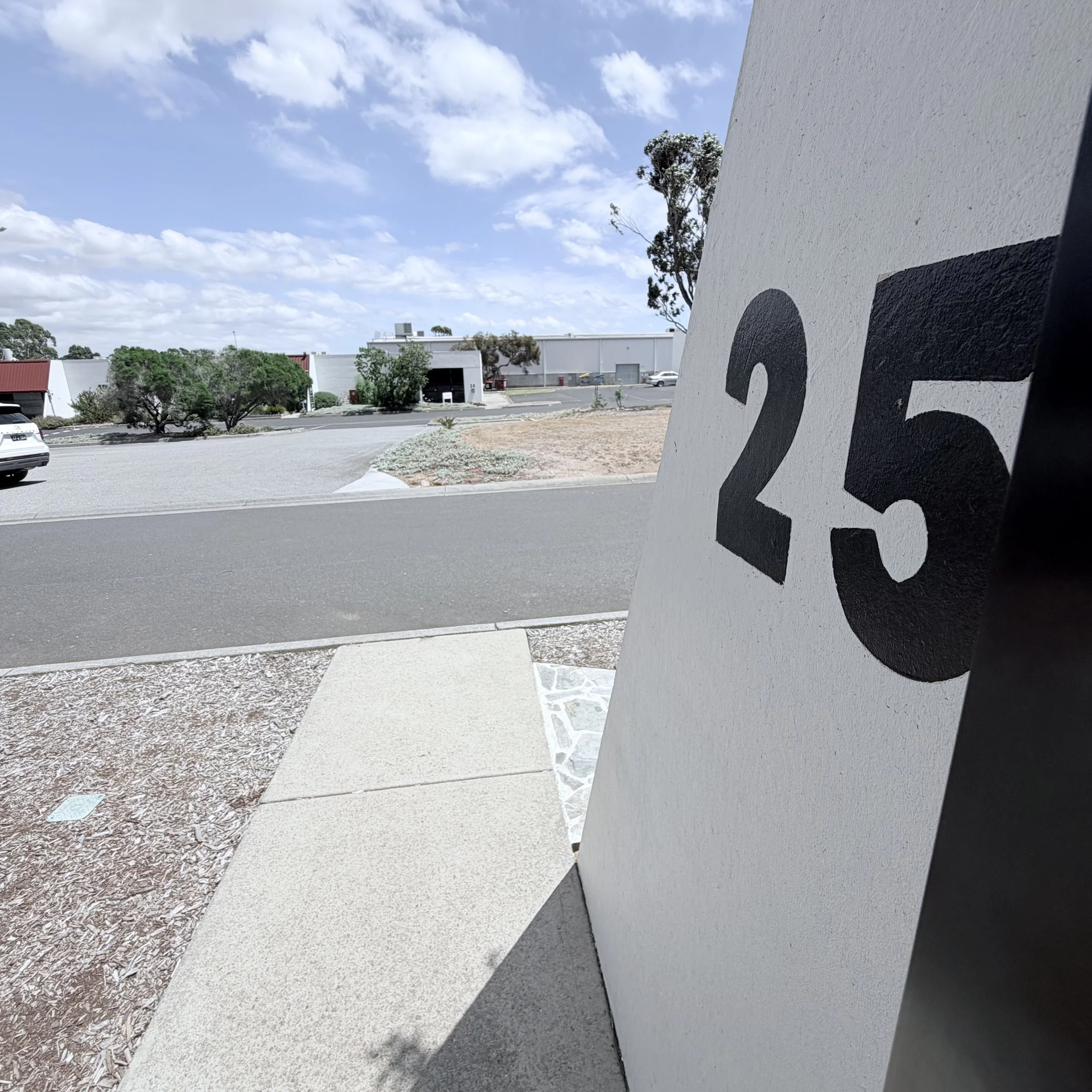A parking lot with some buildings, trees, cars, and a clear sky with scattered clouds. Part of a large painted sign with the letter 'G' on a white wall in the foreground.