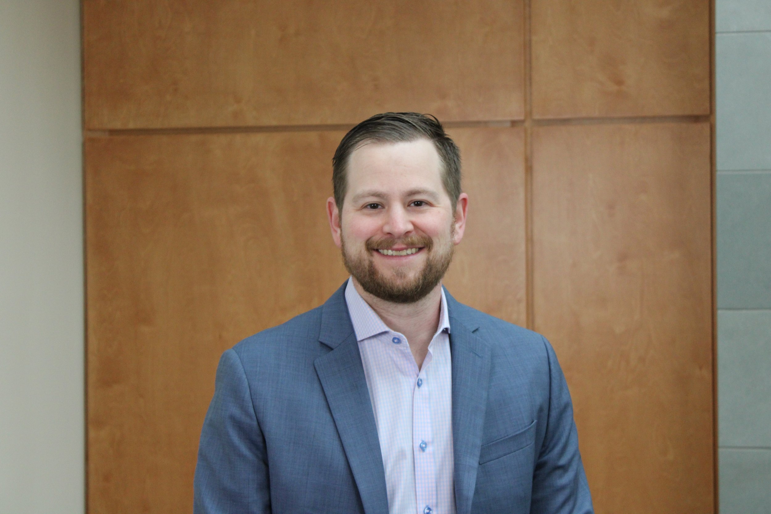 A young man with a beard and short dark hair, dressed in a blue suit and light shirt, standing indoors in front of a wooden wall and smiling at the camera.
