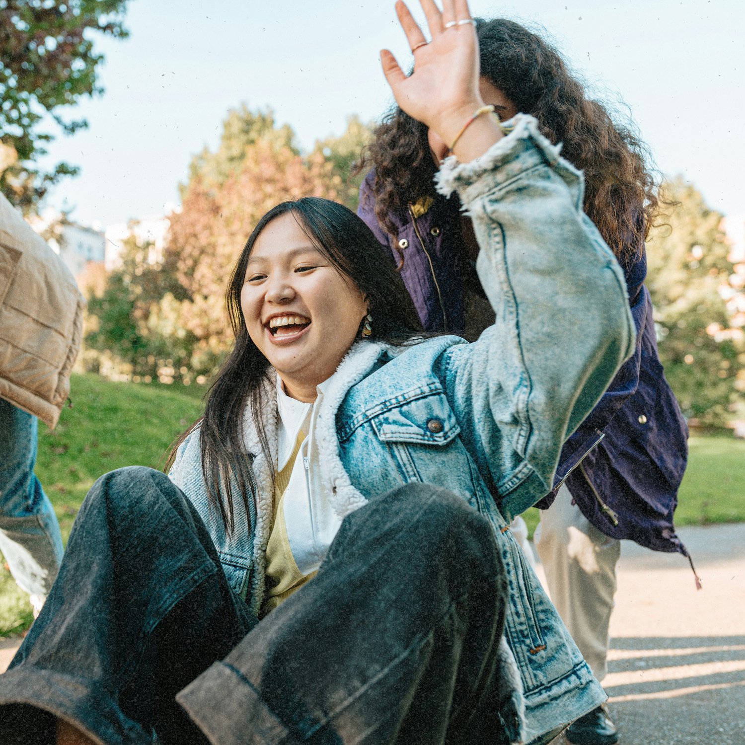 Two young women laughing and smiling outdoors in a park with autumn trees in the background. One woman, with black hair, is sitting down, wearing a denim jacket and jeans, while the other, with curly hair, is standing and partially covering her face with her hand.