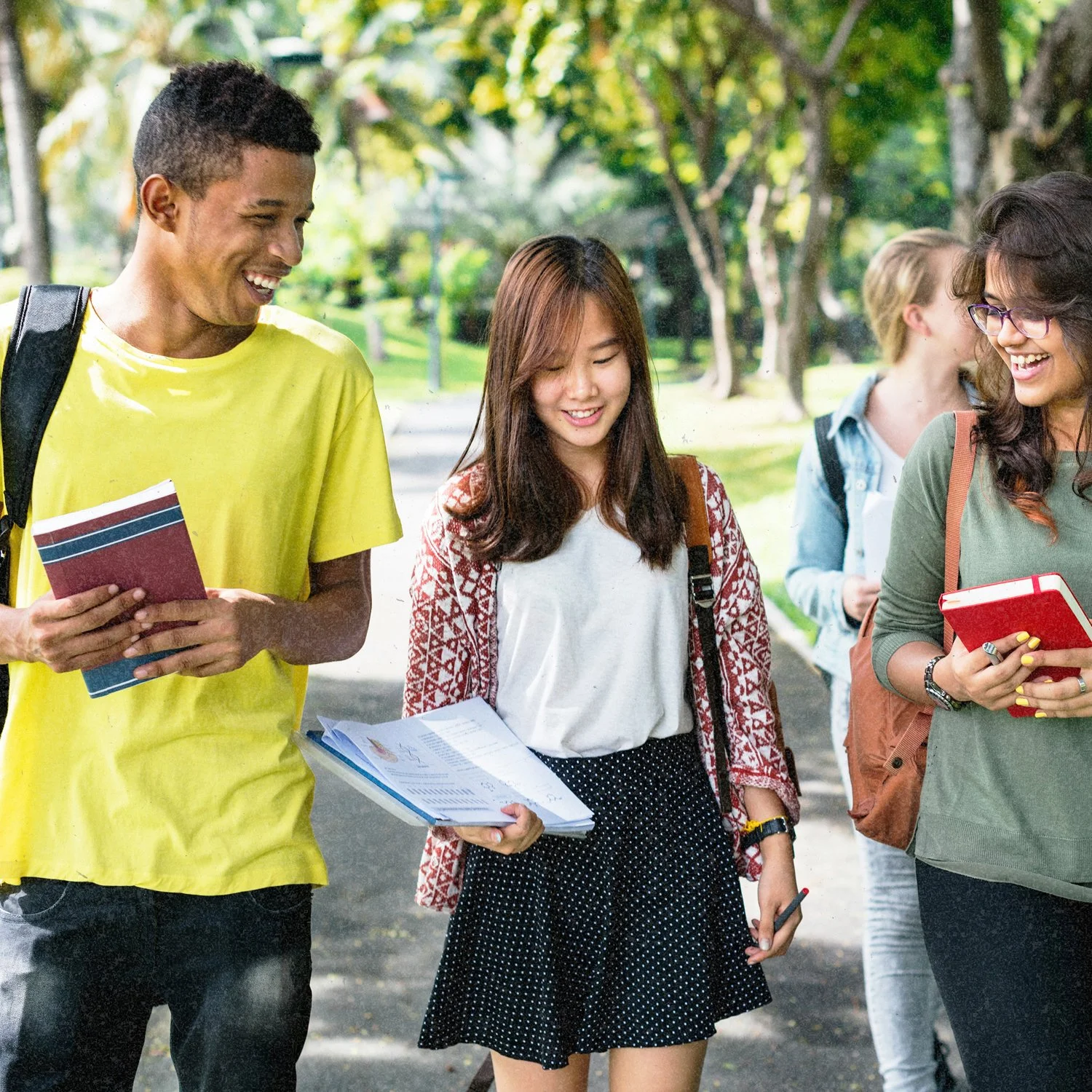Group of four diverse young students chatting outdoors in a park, holding notebooks and papers, with trees and green foliage in the background.