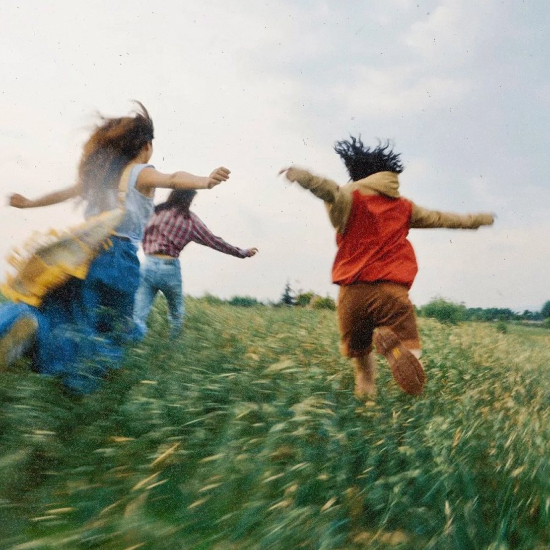 Three teenagers running through a green field on a cloudy day.
