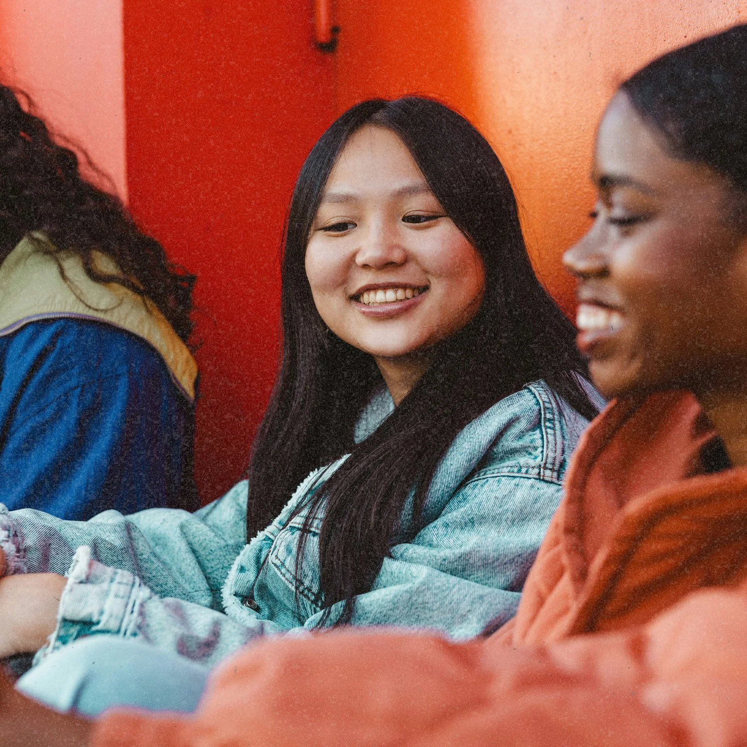 Three young women sitting together, engaging in conversation, with one of them smiling at the woman in the center, in a room with a red/orange background.