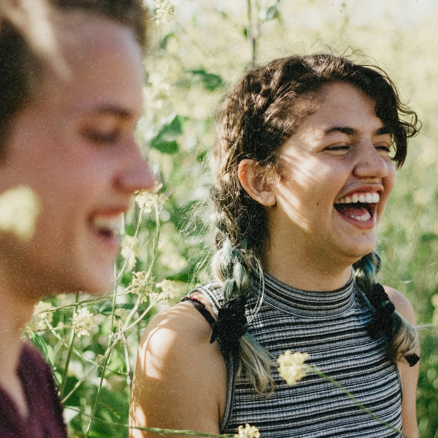 Two teeangers smiling in a green outdoor setting, one with dark curly hair and the other with light brown hair, surrounded by wildflowers.