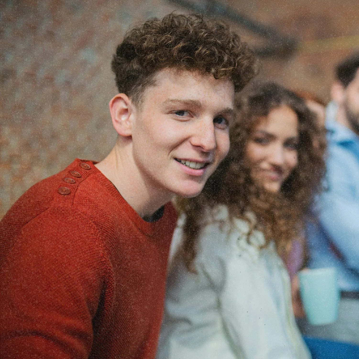A young man with curly hair wearing a red sweater smiling, sitting next to a woman with curly hair wearing a white jacket, in a social setting.
