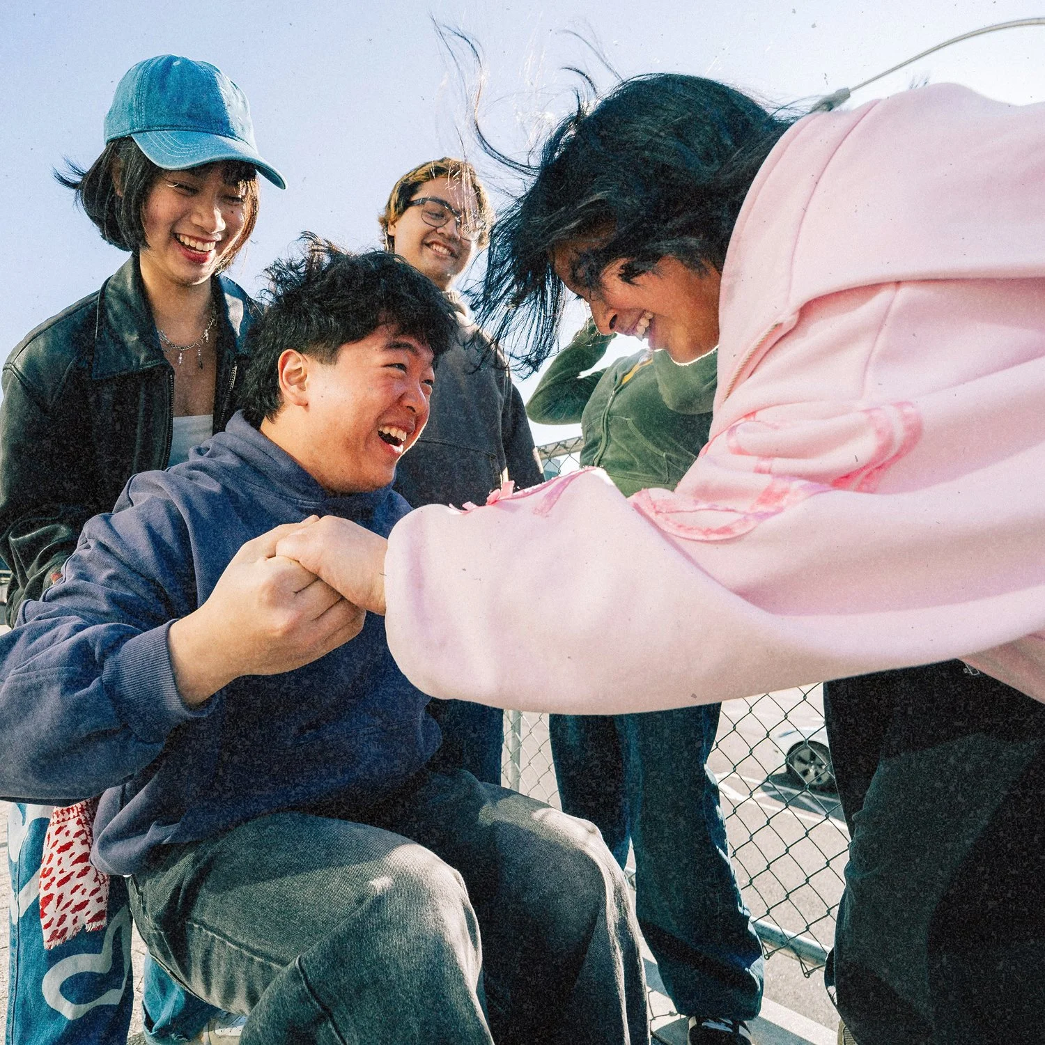 A group of five friends is outside, smiling and enjoying themselves. Two of them are holding hands, with woman in a pink hoodie engaged in the interaction. The group is casually dressed, and the overall mood is joyful and friendly.