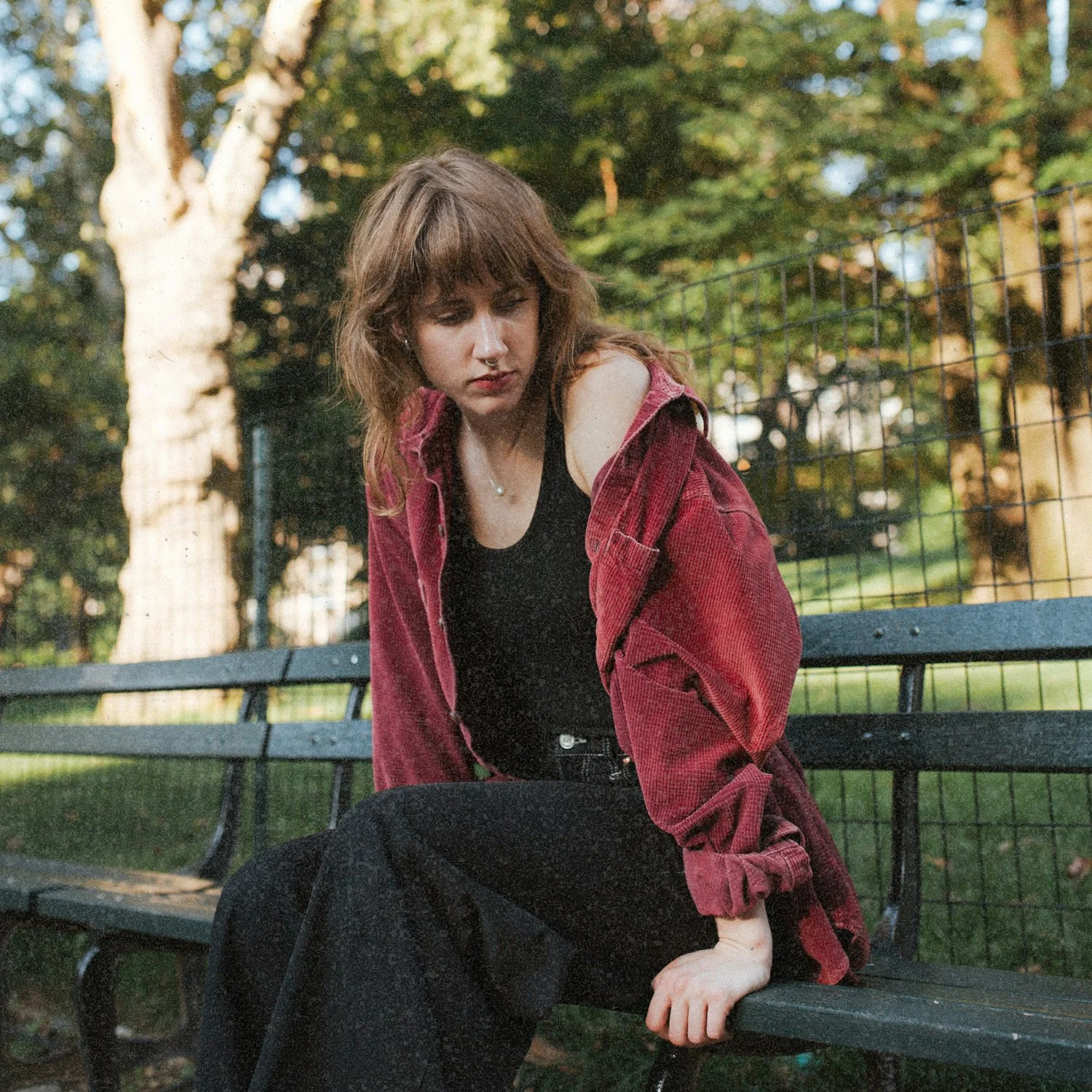 A young woman with wavy, shoulder-length brown hair sits on a school bench, wearing a pink jacket and black pants, looking down with a contemplative expression.