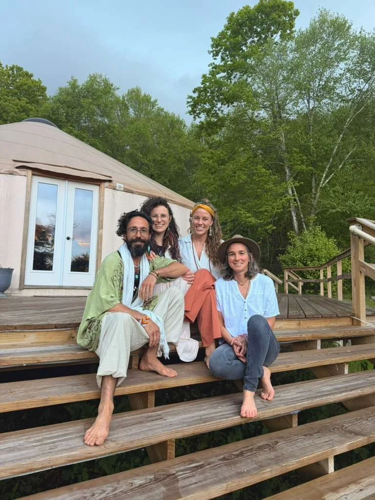 Four people sitting together on wooden steps in front of a house with trees in the background, smiling at the camera, enjoying a relaxed outdoor moment.