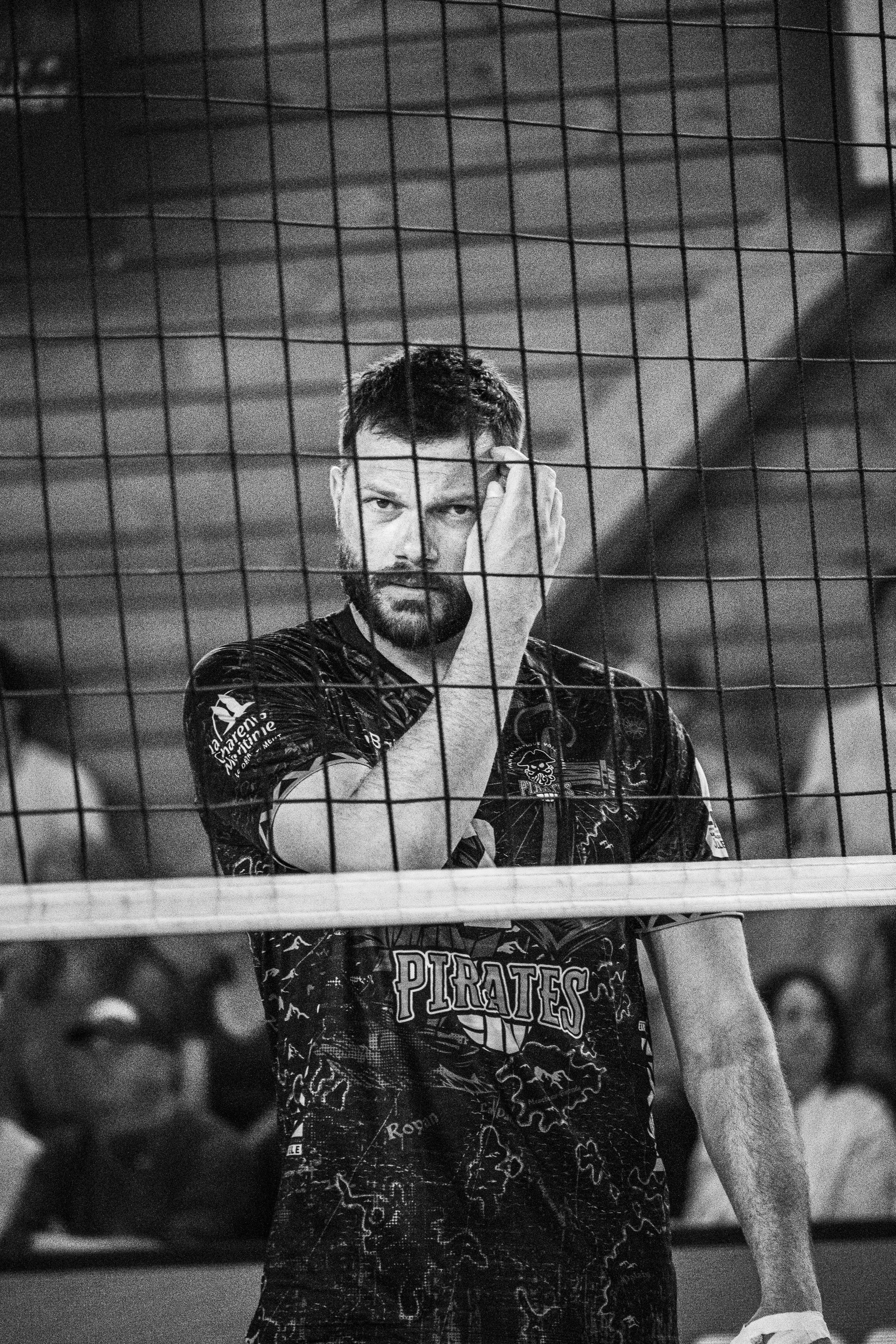 Un homme portant un maillot de volleyball avec le logo 'Pirates', une barbe et une coiffure courte, tate la tête avec sa main, derrière une grille de volleyball.